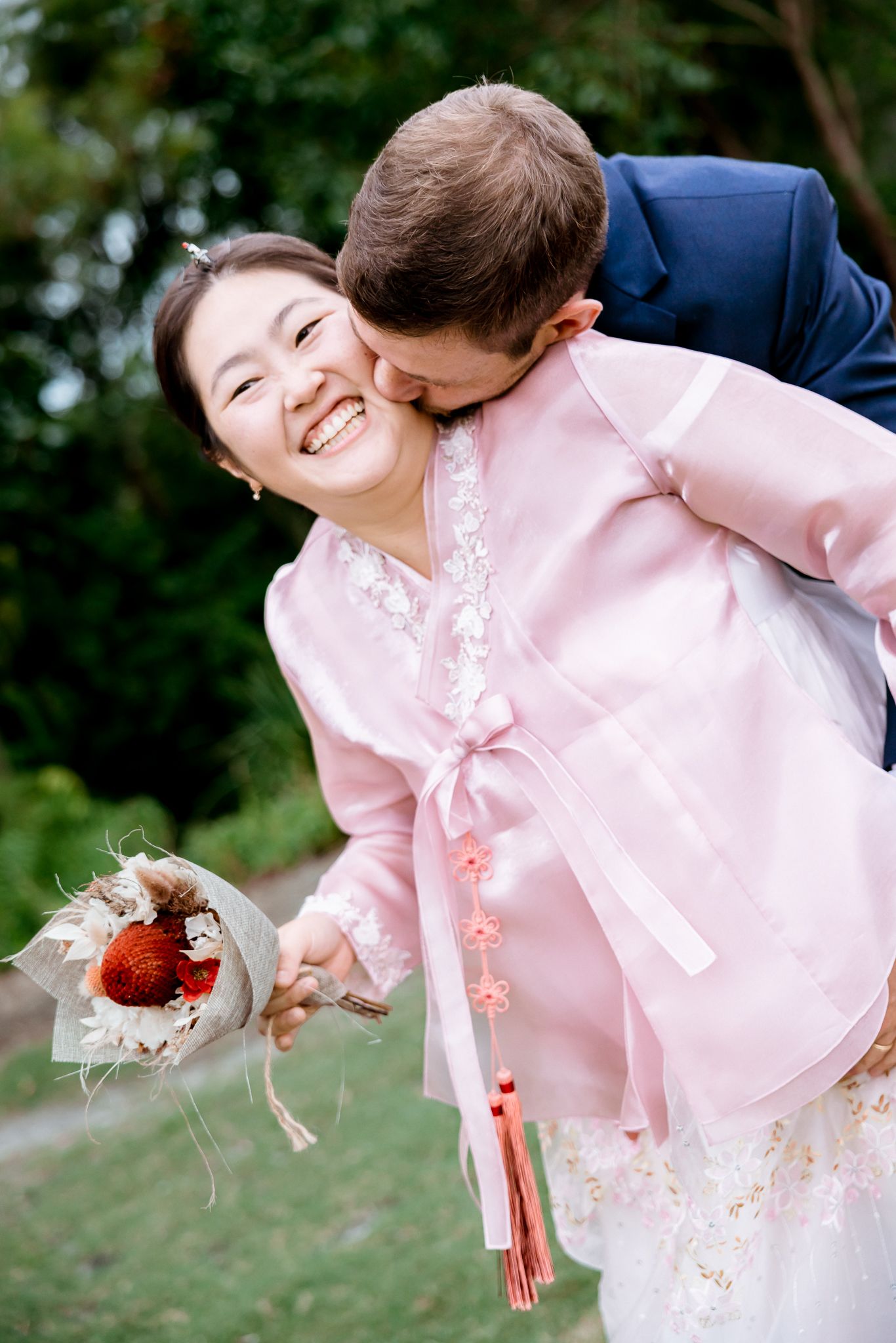 Smiling bride in pink hanbok holds bouquet while groom in blue suit kisses her cheek outdoors.