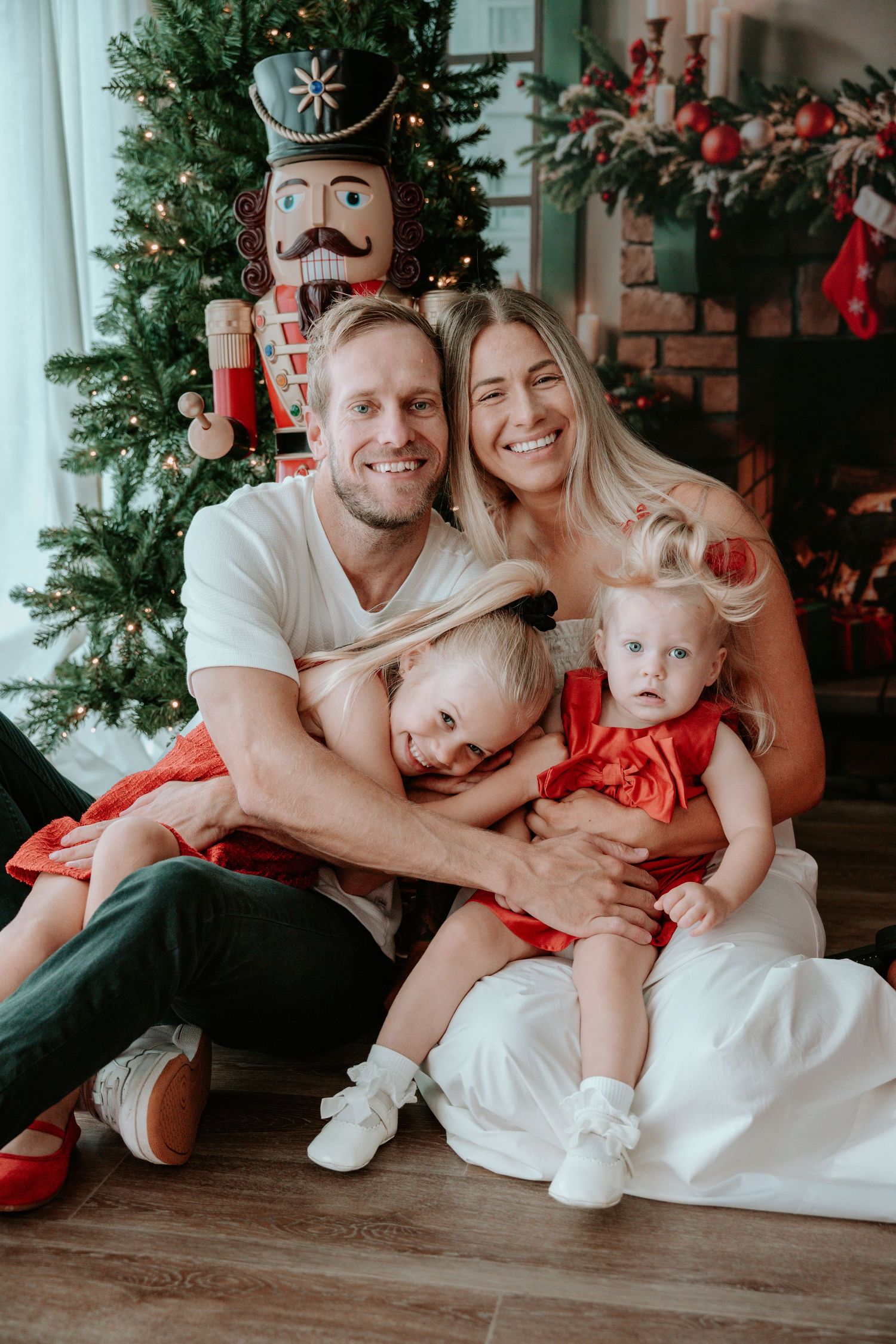 Happy family of four posing by decorated Christmas tree with nutcracker, parents hugging two young daughters in red dresses.