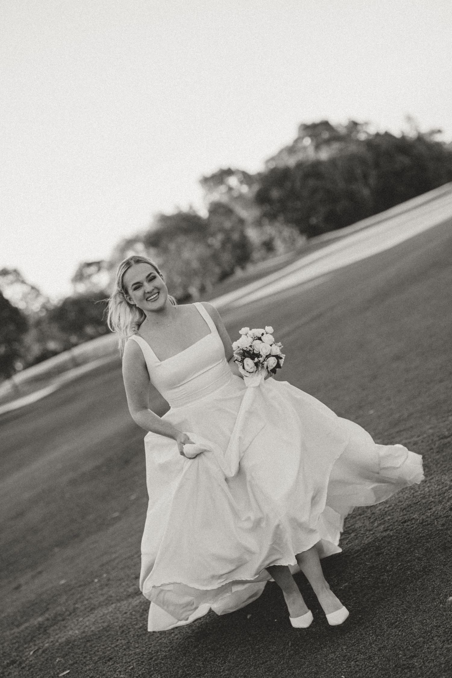 Bride in white dress twirling on grassy hill in vintage black and white style.