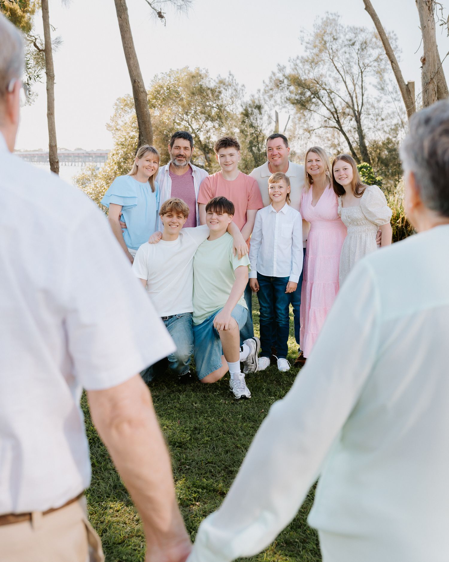 Large family group photo outdoors on green lawn surrounded by trees, wearing pastel outfits, smiling together.