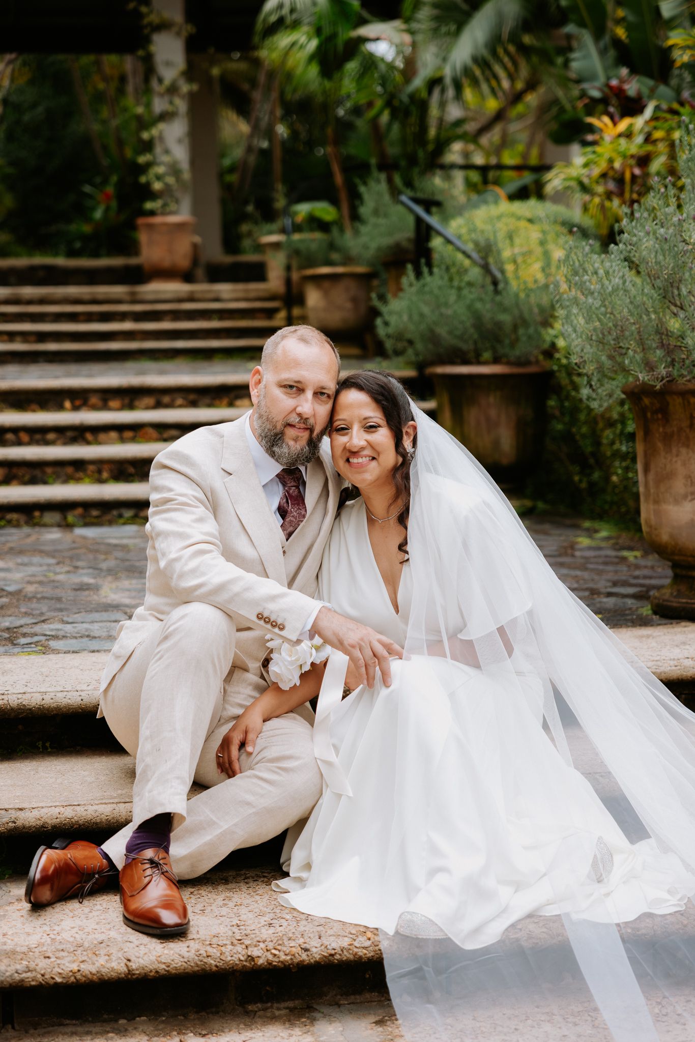 A bride and groom sit together on stone steps surrounded by lush greenery, smiling warmly on their wedding day.