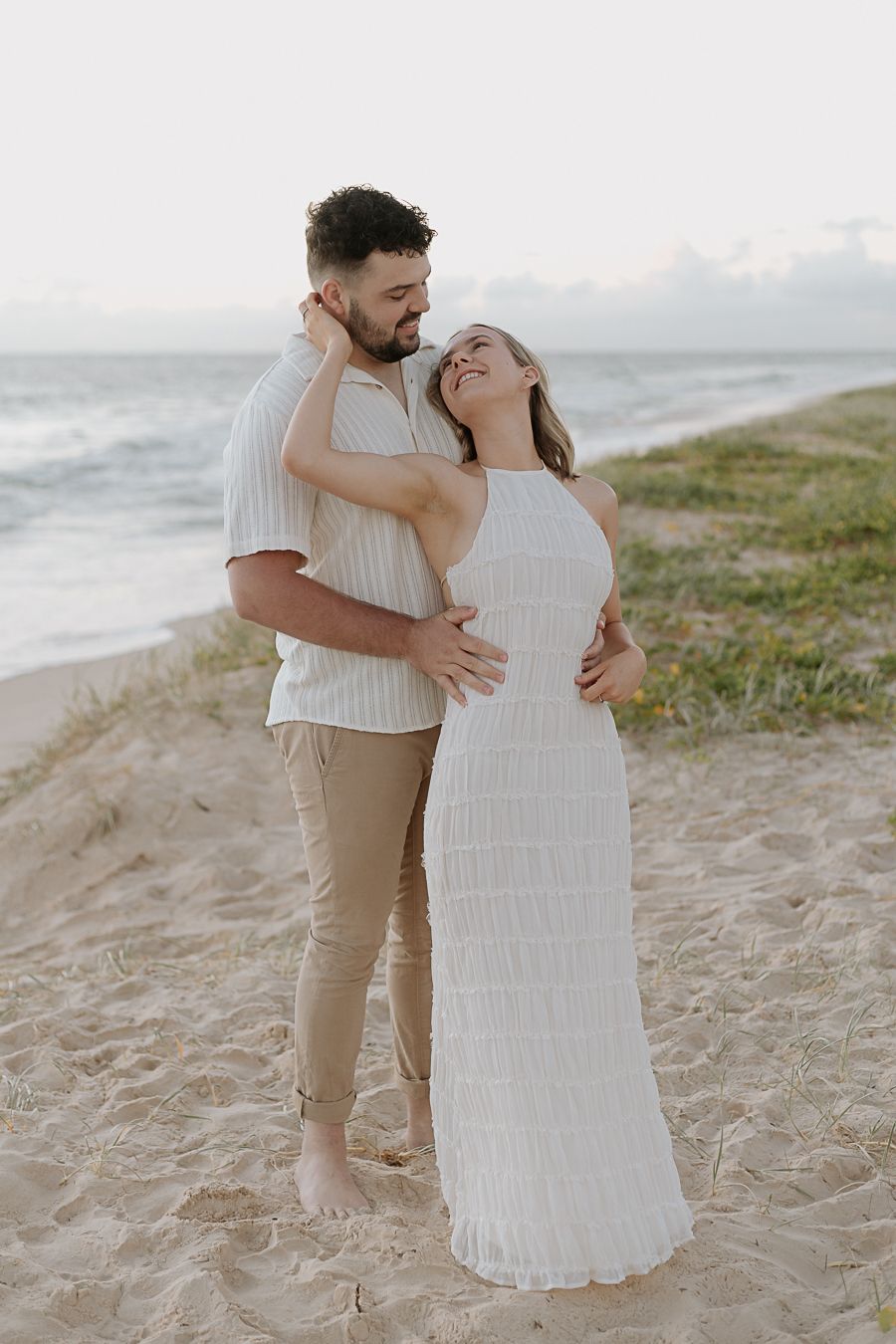 Couple embracing on sandy beach at sunset, woman in white dress smiling up at bearded man in striped shirt.