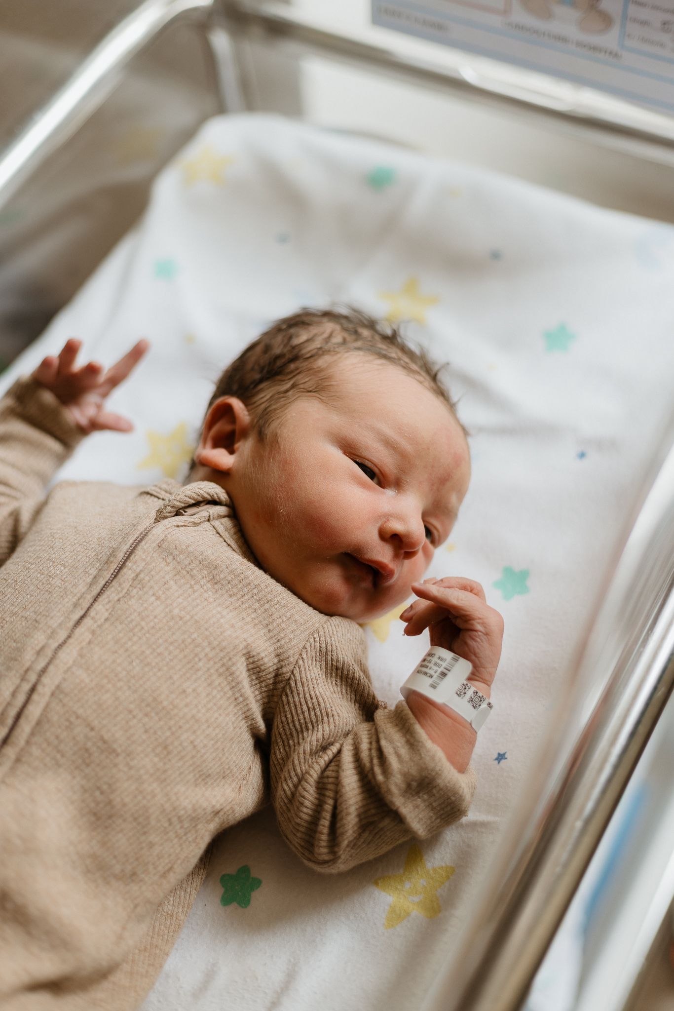 A sleeping newborn wearing a beige sweater lies in a hospital bassinet with colorful star decorations.