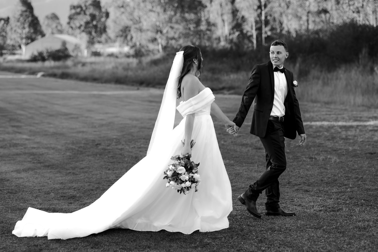 Black and white photo of bride and groom holding hands walking across grass at outdoor wedding.