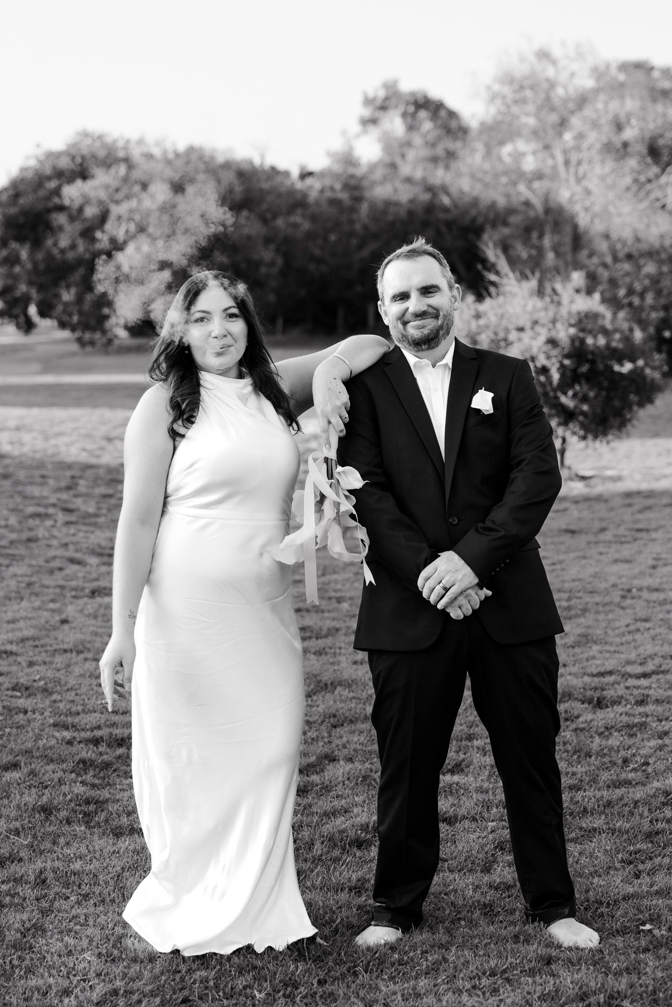 Black and white wedding photo of bride in white gown and groom in suit smiling outdoors on grass.