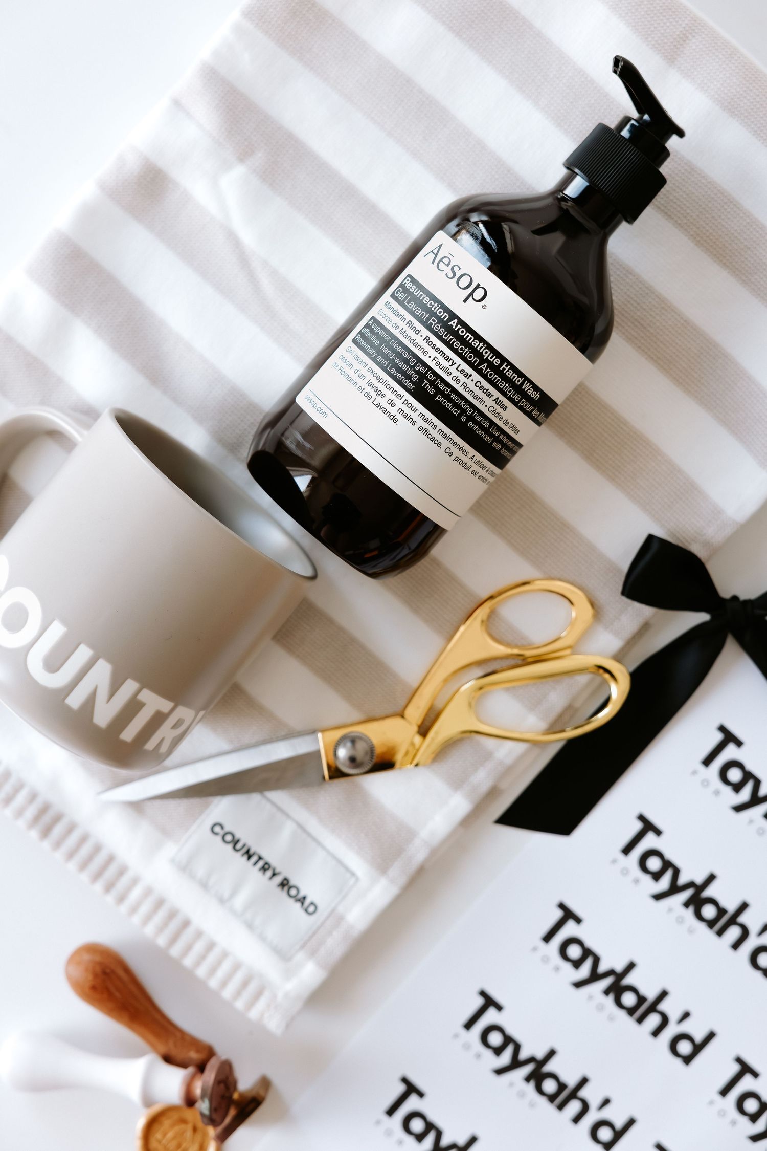 Flat lay of Aesop hand soap, gold scissors, Country Road mug, striped towel, and gift tags with black ribbon.