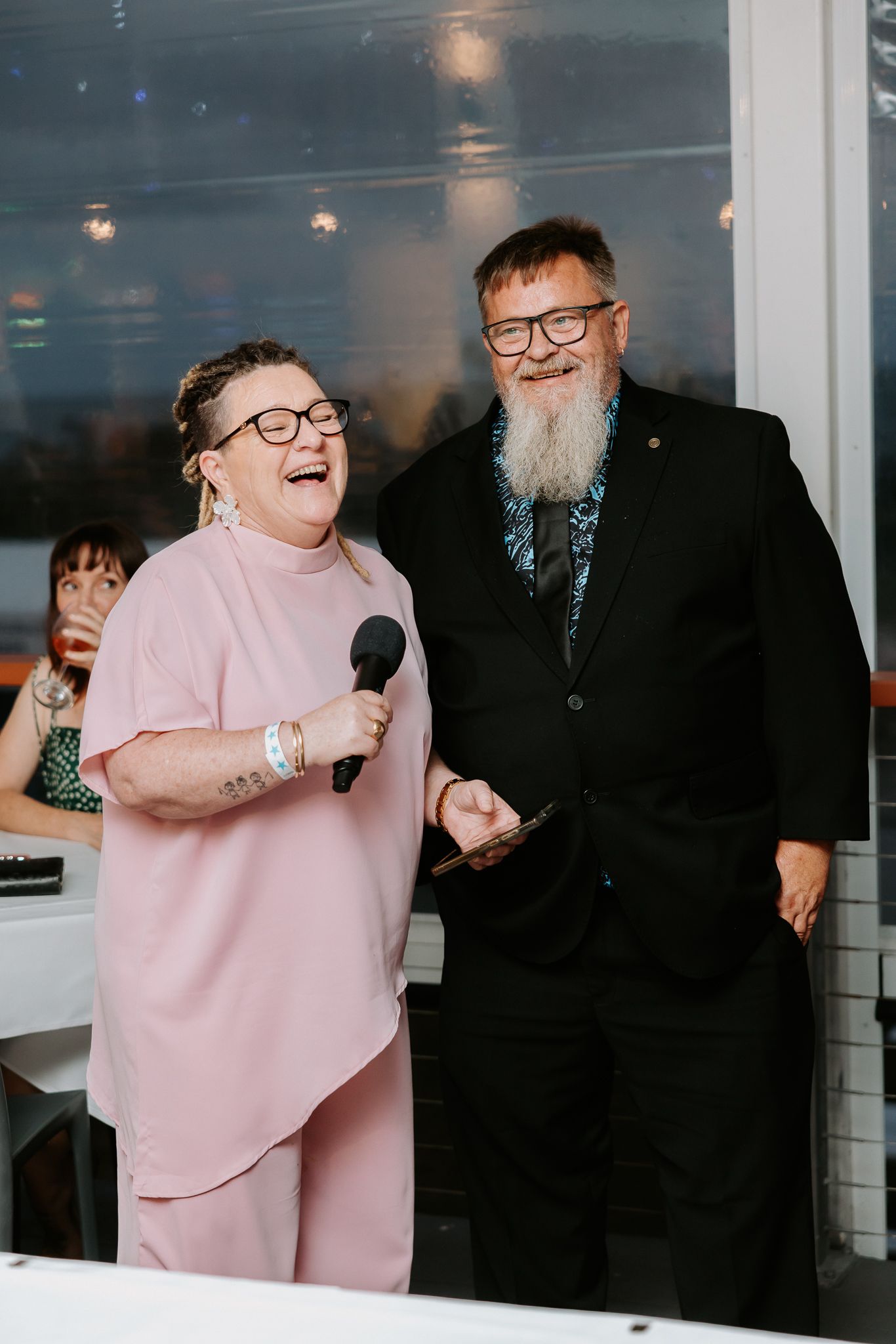 Woman in pink suit speaking into microphone beside bearded man in black suit at wedding reception.