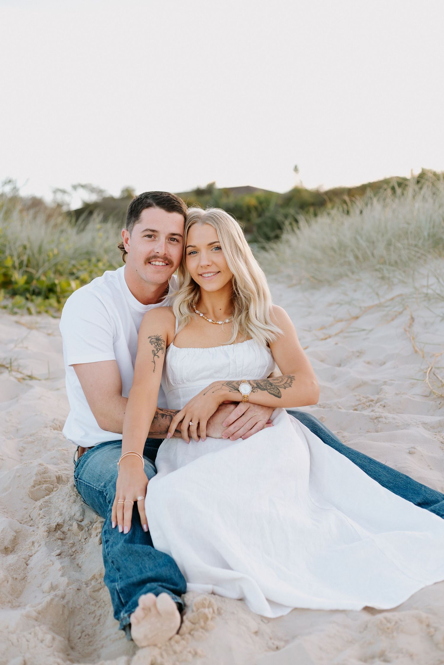 A couple sits together on sandy beach dunes, woman in white dress and man in white tee and jeans, smiling warmly.