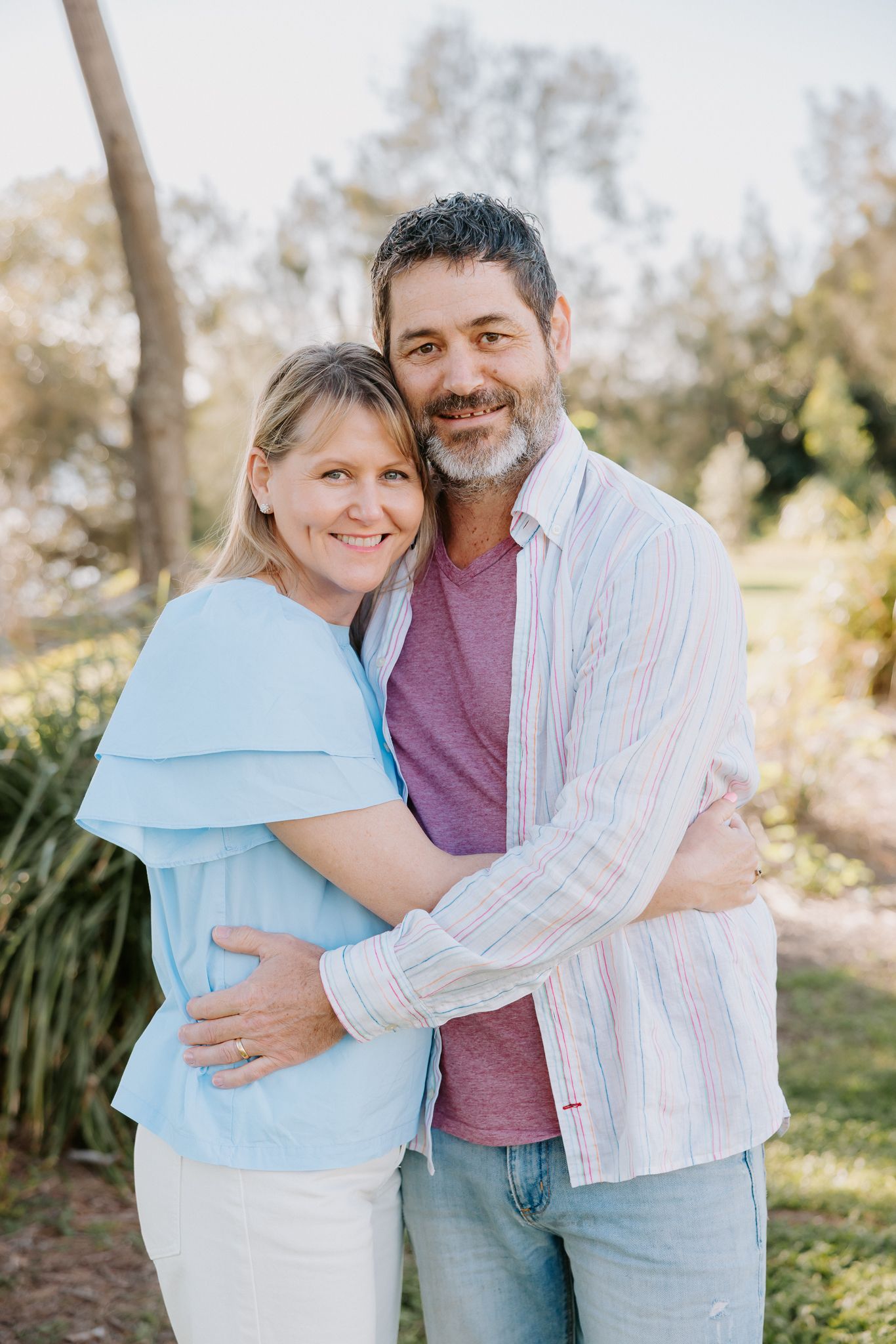 A smiling middle-aged couple embracing outdoors in a park setting, woman in light blue top and man in striped shirt.
