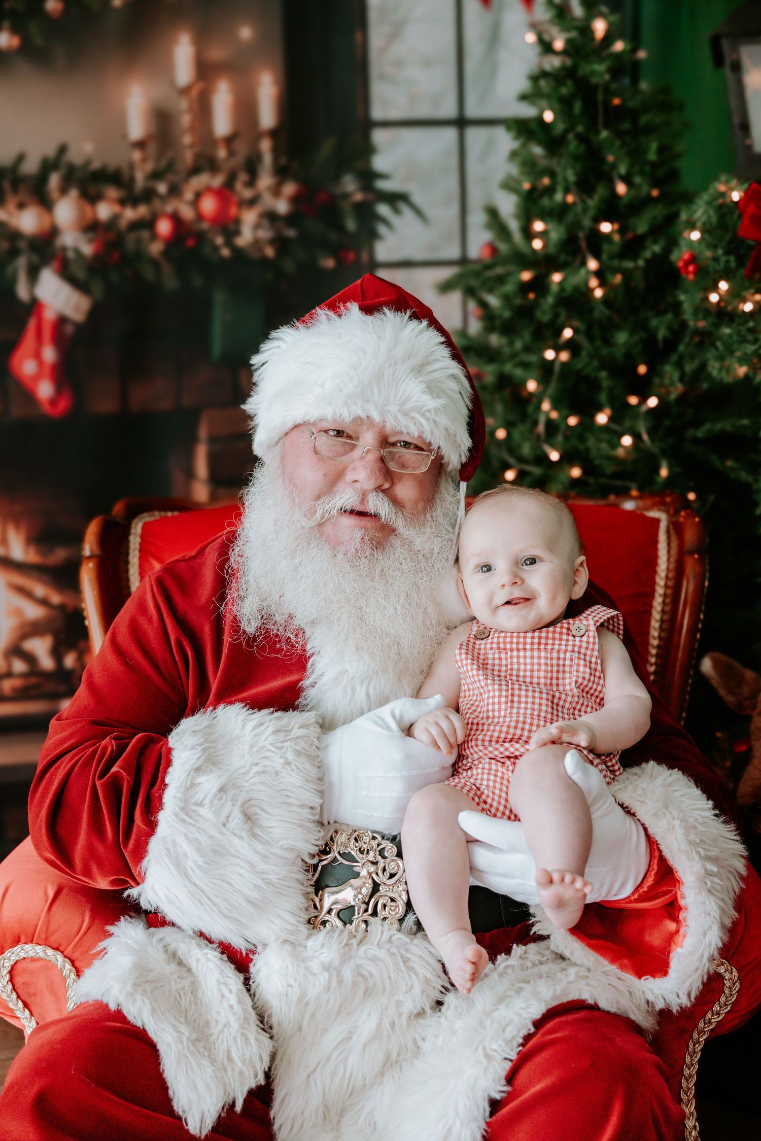 Baby girl in red dress sits on Santa's lap in front of a decorated Christmas tree and fireplace.