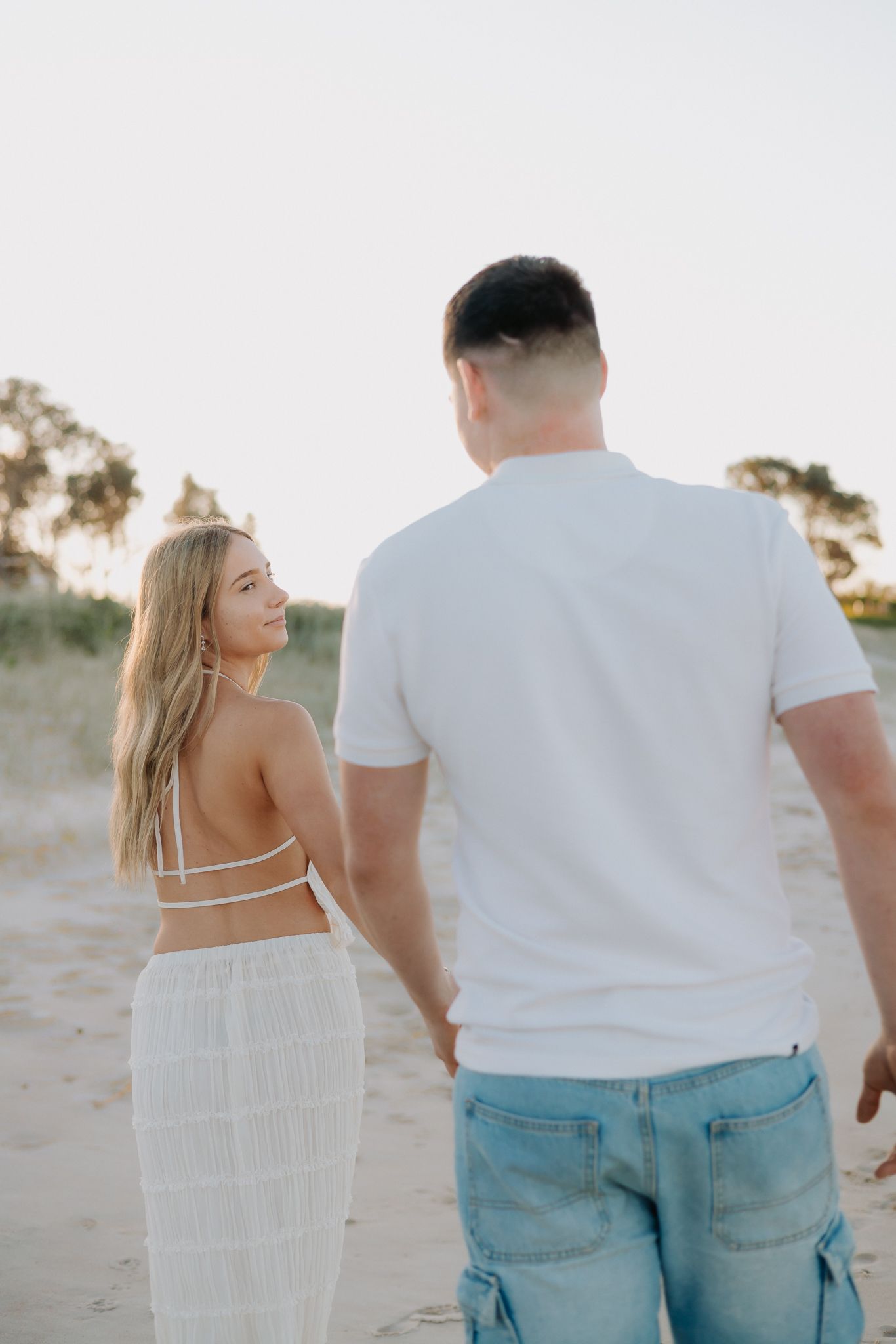 Couple holding hands walking on sandy beach at sunset, woman in white backless dress glances back smiling.