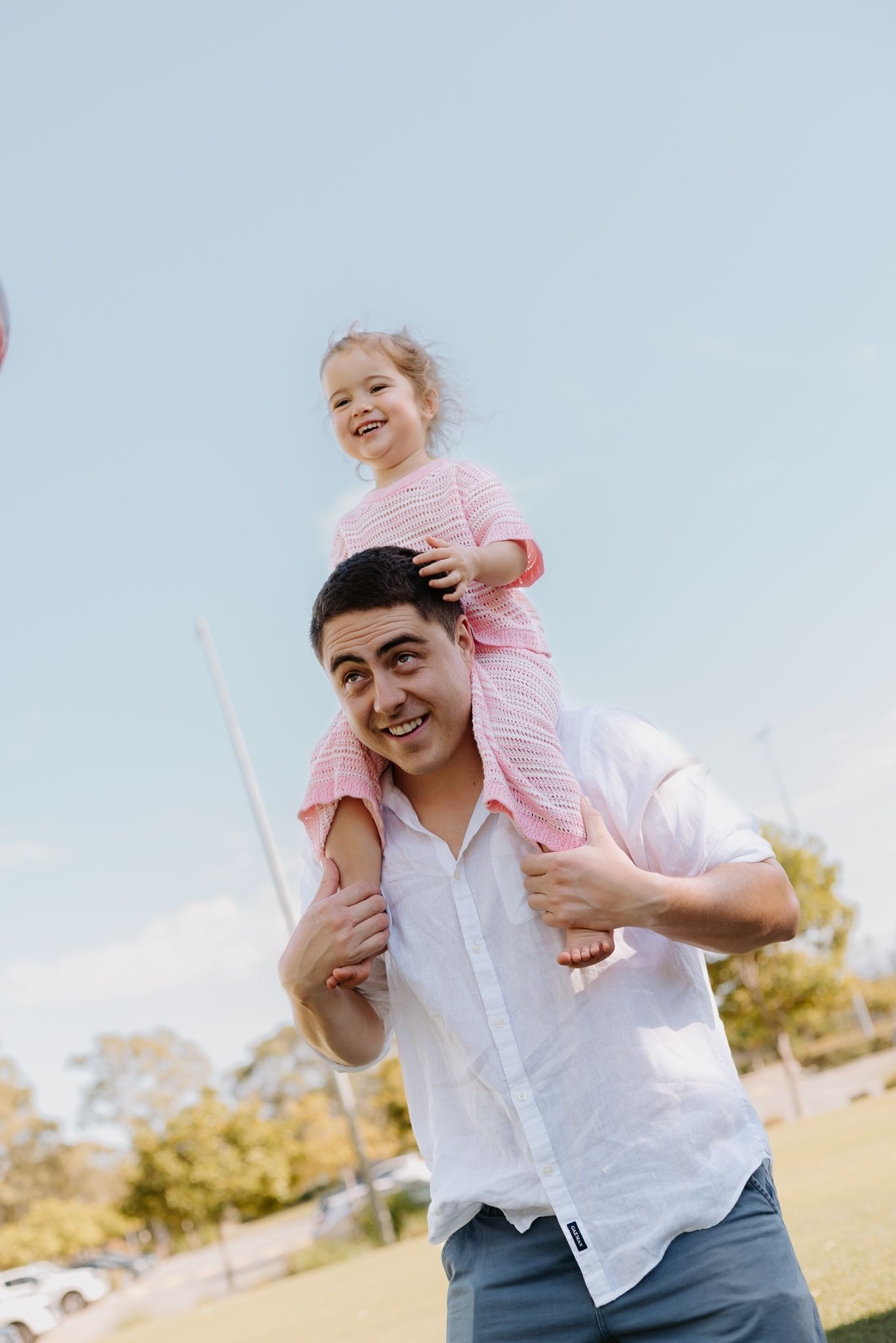 A smiling man carries a happy young girl on his shoulders outdoors in a sunny park setting.