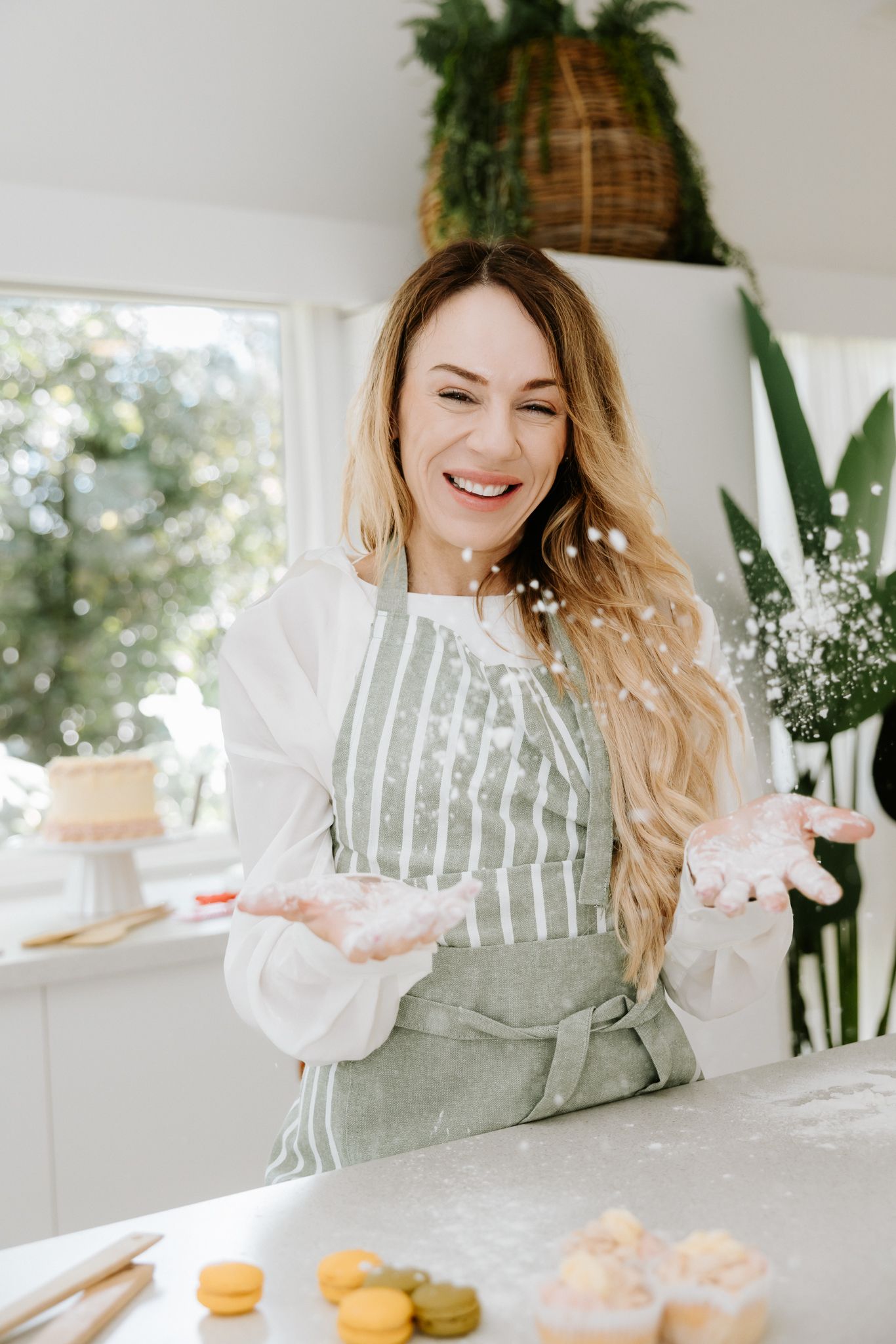 Smiling woman in striped apron clapping flour in bright kitchen with macarons and cake on counter.