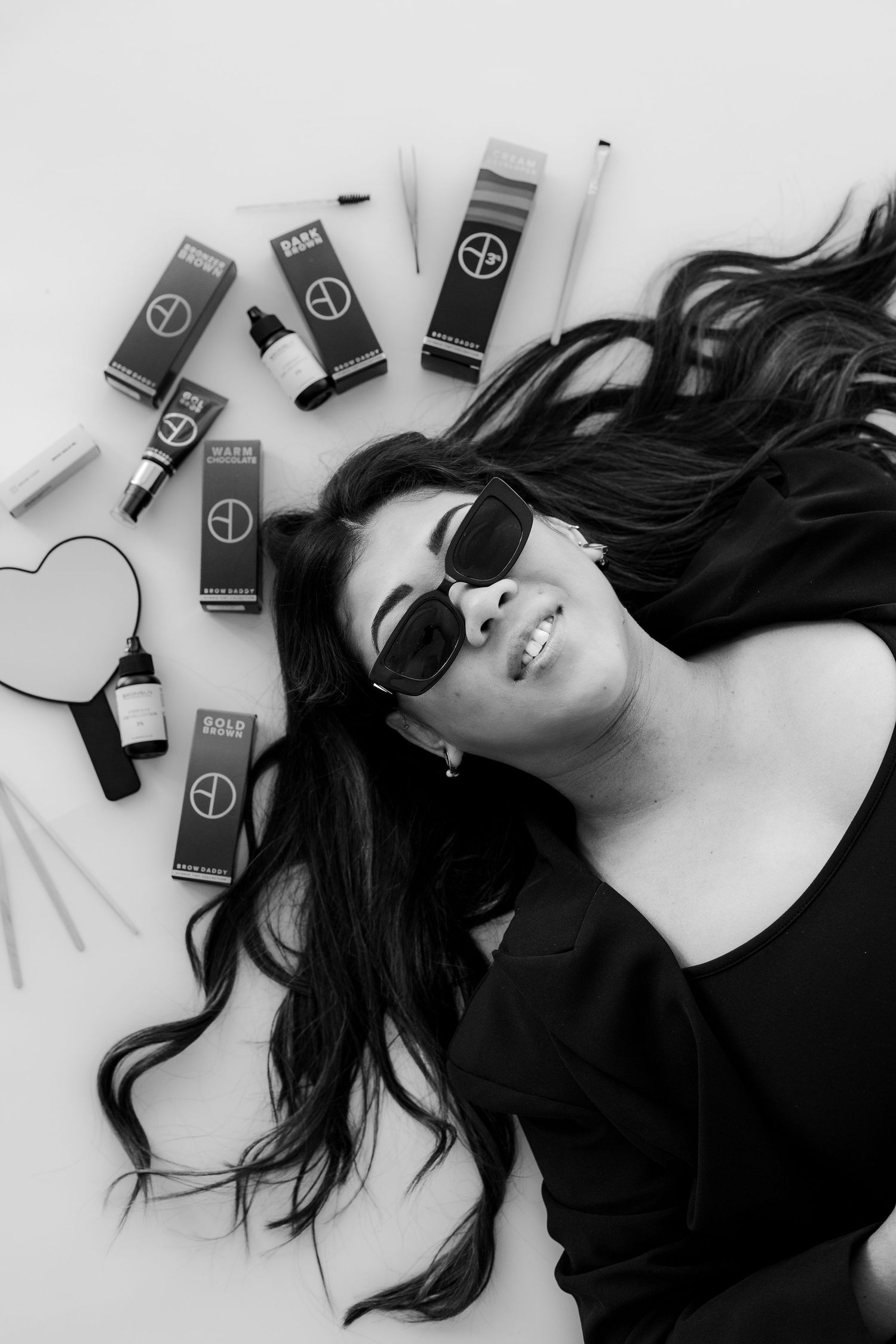 Black and white photo of a smiling woman in sunglasses lying among beauty products with long flowing hair.
