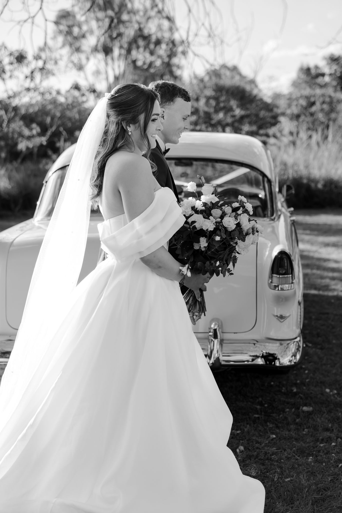 Black and white photo of bride in flowing gown with veil and bouquet standing with groom beside vintage car.