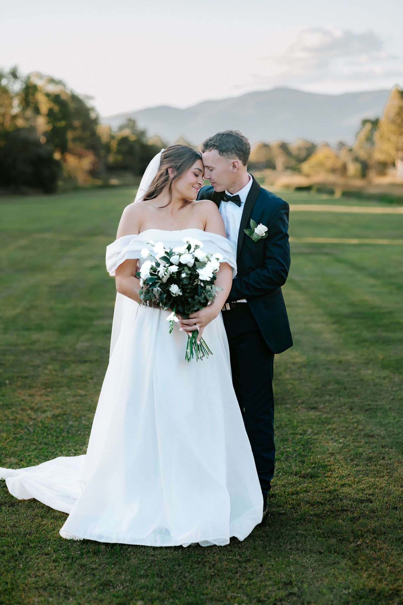 Bride and groom share a romantic moment on green lawn with mountain backdrop, bride holding white floral bouquet.