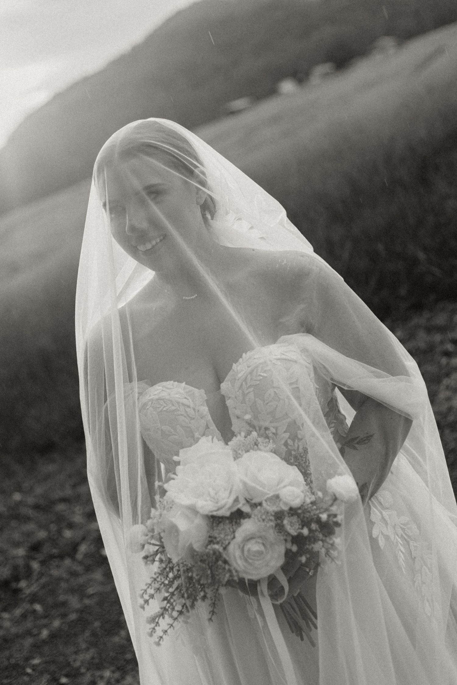 Ethereal black and white bridal portrait with flowing veil and white rose bouquet against misty outdoor backdrop.