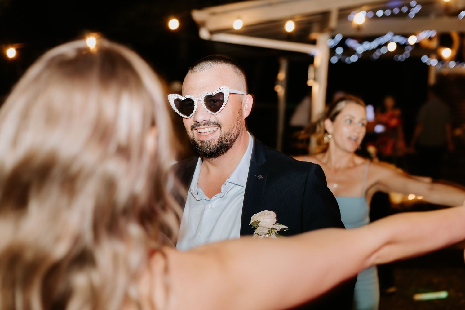 Wedding guest in sunglasses and suit jacket smiling during outdoor evening reception celebration.