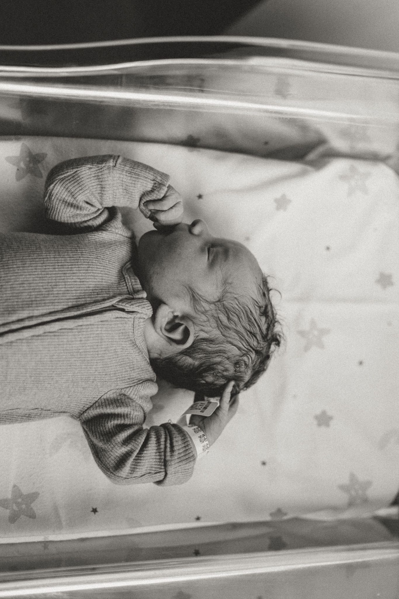 Black and white photograph of a newborn baby sleeping in a hospital bassinet on star-patterned bedding.