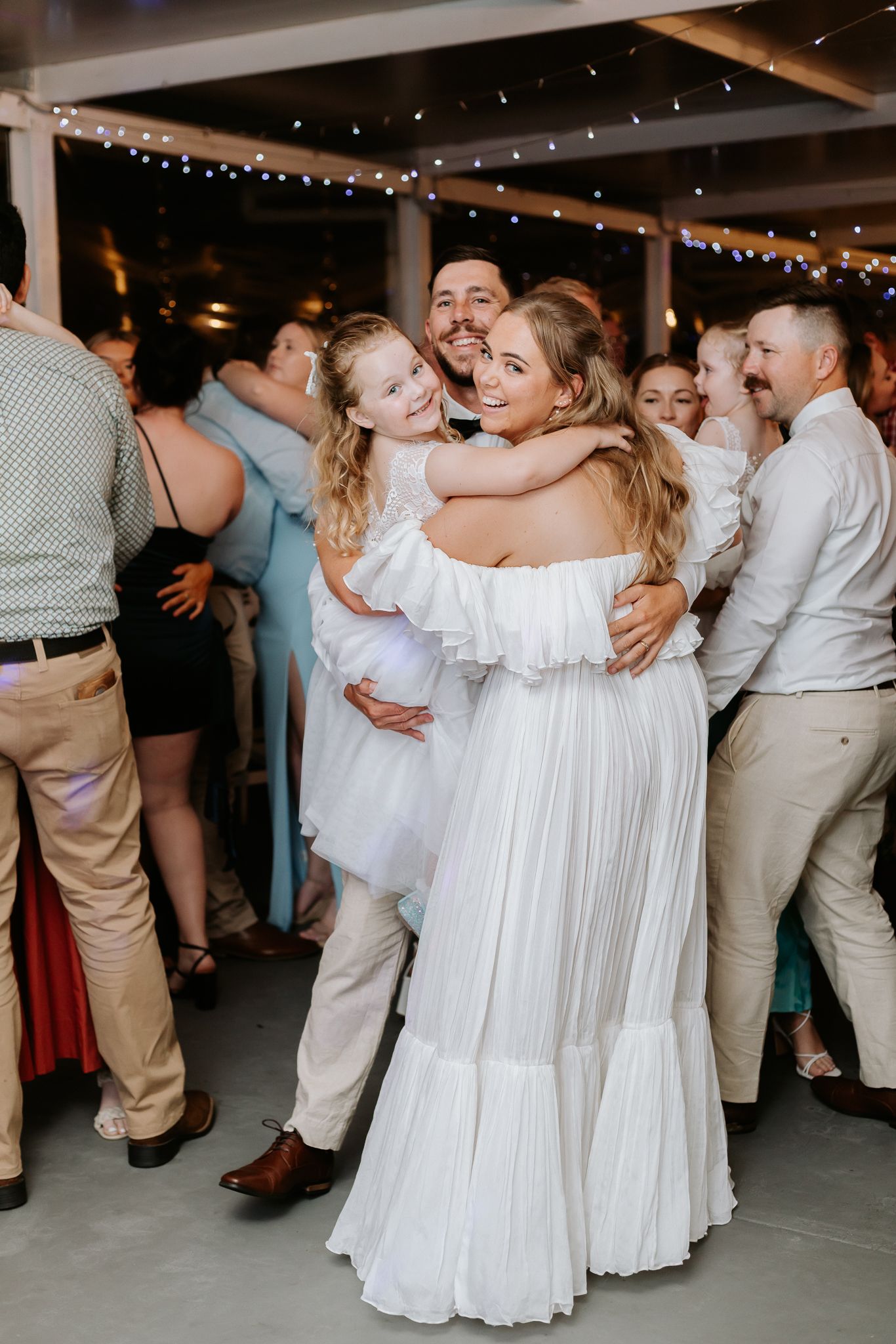 Bride and groom hugging a young flower girl on the dance floor surrounded by wedding guests.