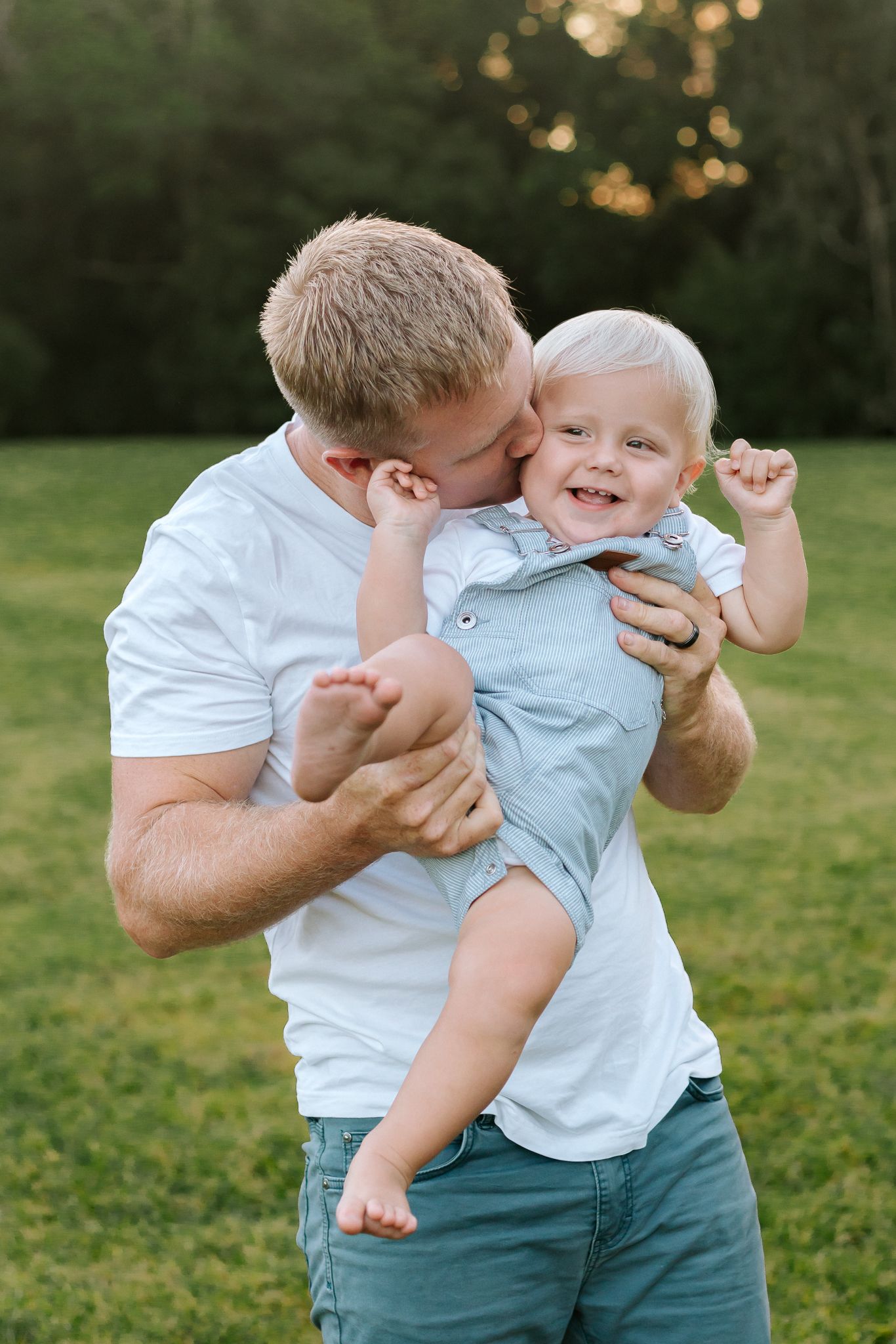 Father kissing happy laughing baby boy outdoors in green park during warm sunset.