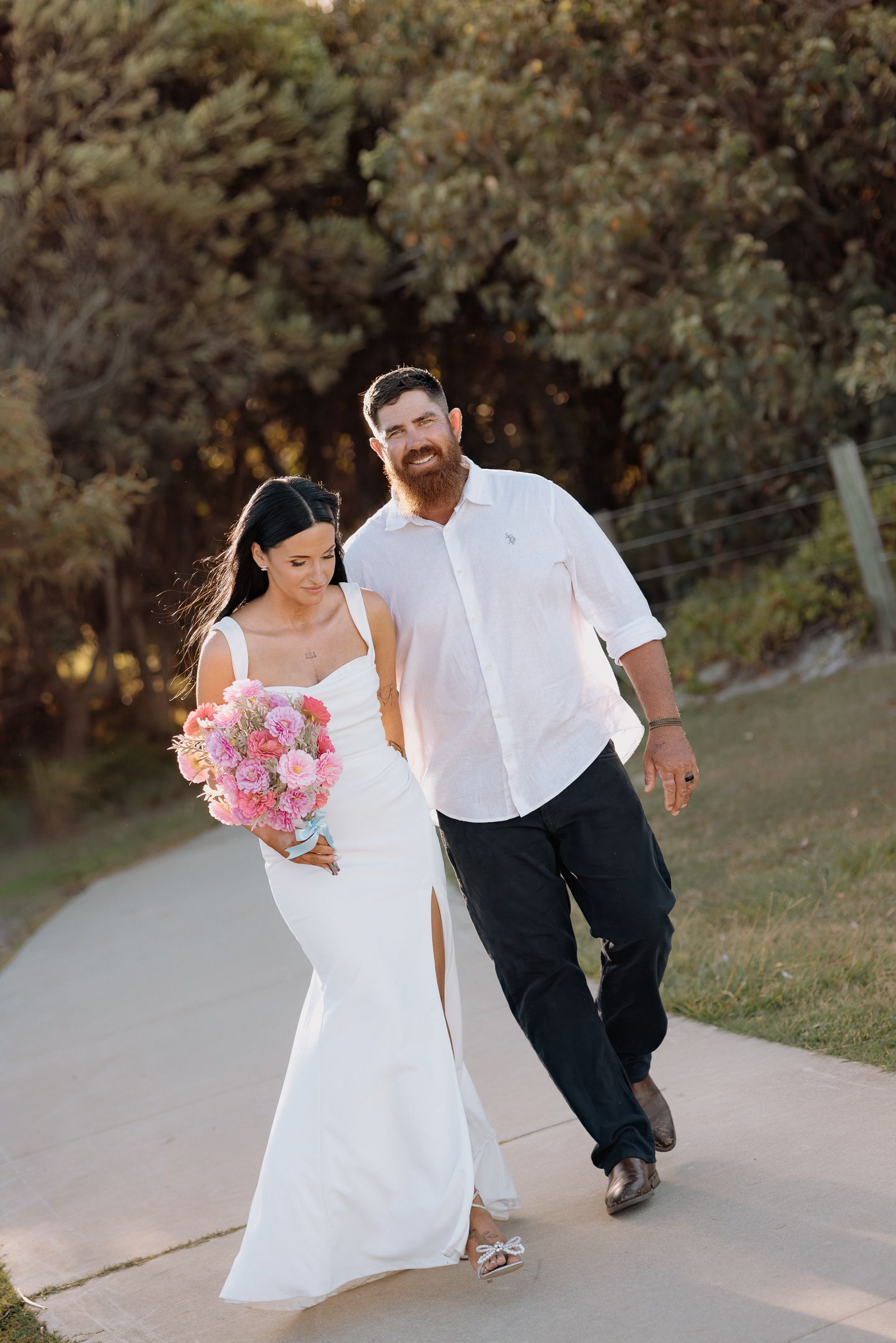 A couple walking together outdoors, with the person in a white dress holding a pink bouquet on a paved path.
