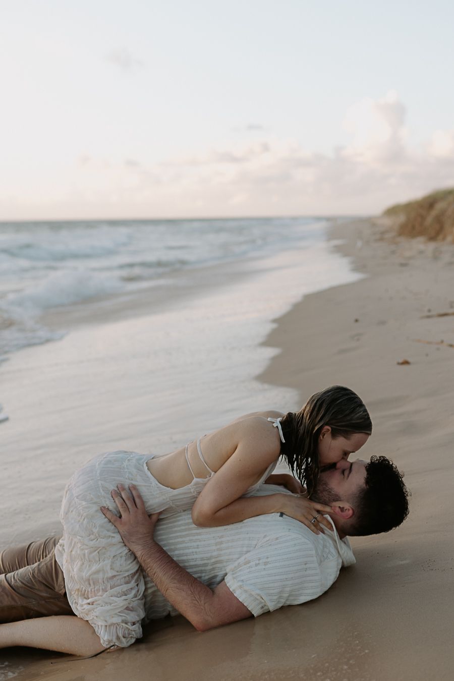 Couple sharing a romantic kiss on a sandy beach at sunset, with ocean waves gently rolling in the background.