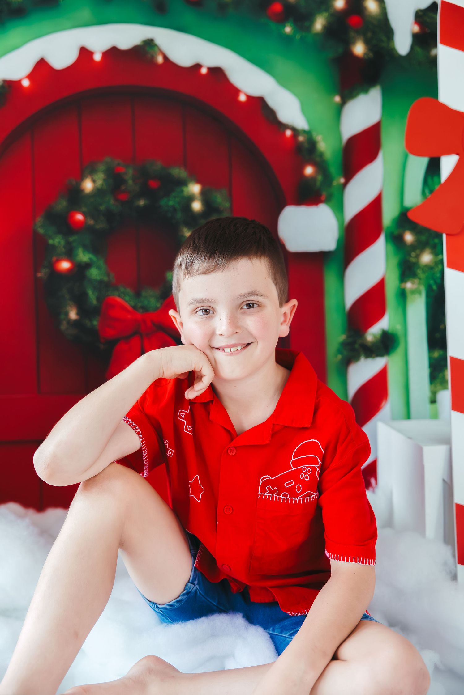 Smiling boy in red shirt sits in front of festive Christmas backdrop with wreath, candy canes, and holiday lights.