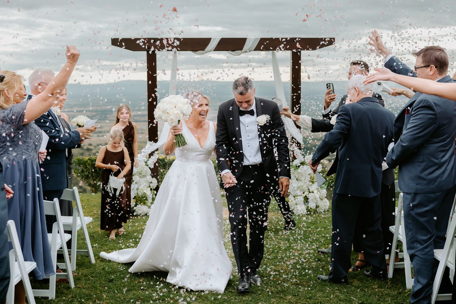 Bride and groom walk down aisle as guests toss white confetti at outdoor wedding ceremony with floral arch backdrop.