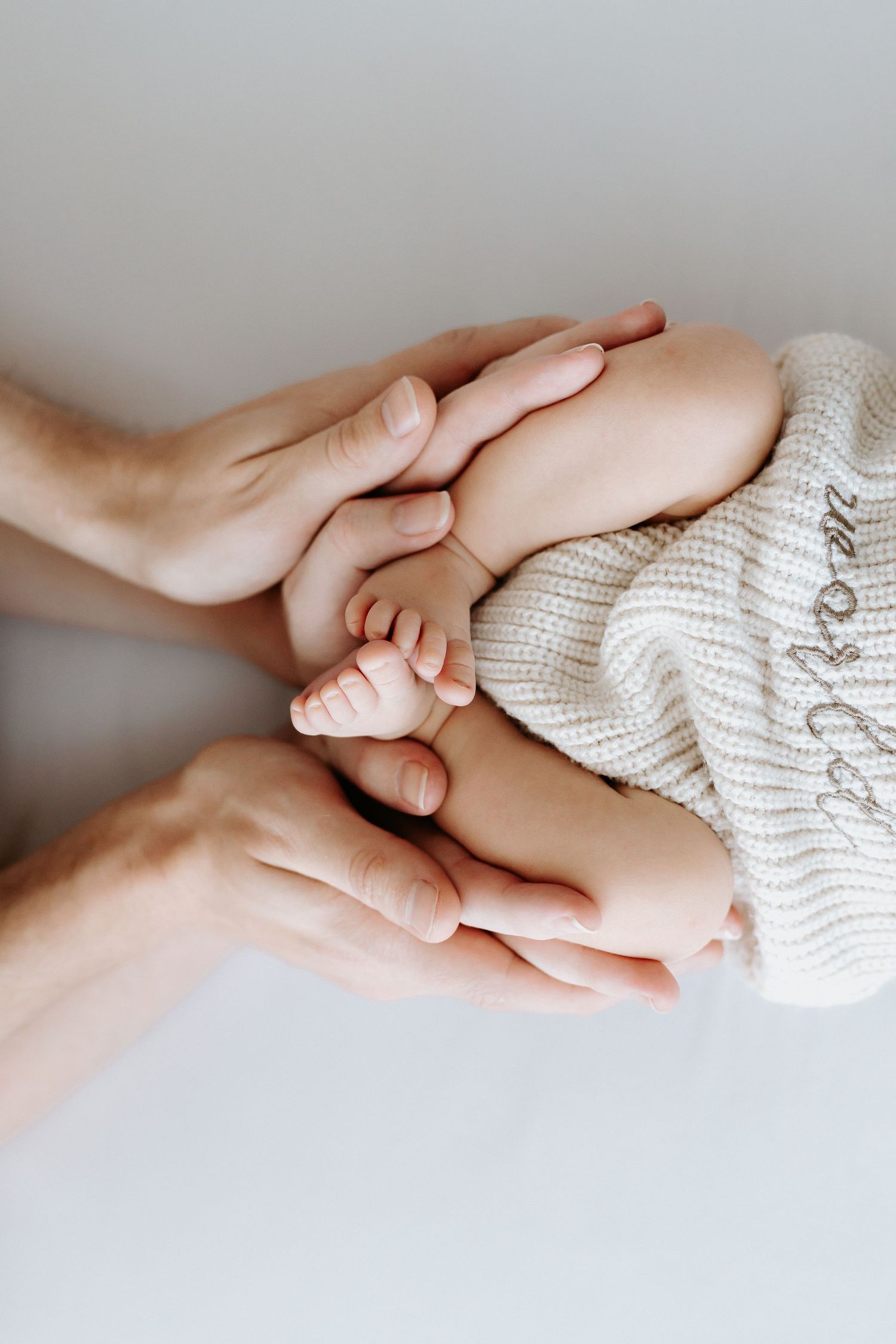 Adult hands gently cradling tiny newborn baby feet wrapped in a soft cream knit blanket.