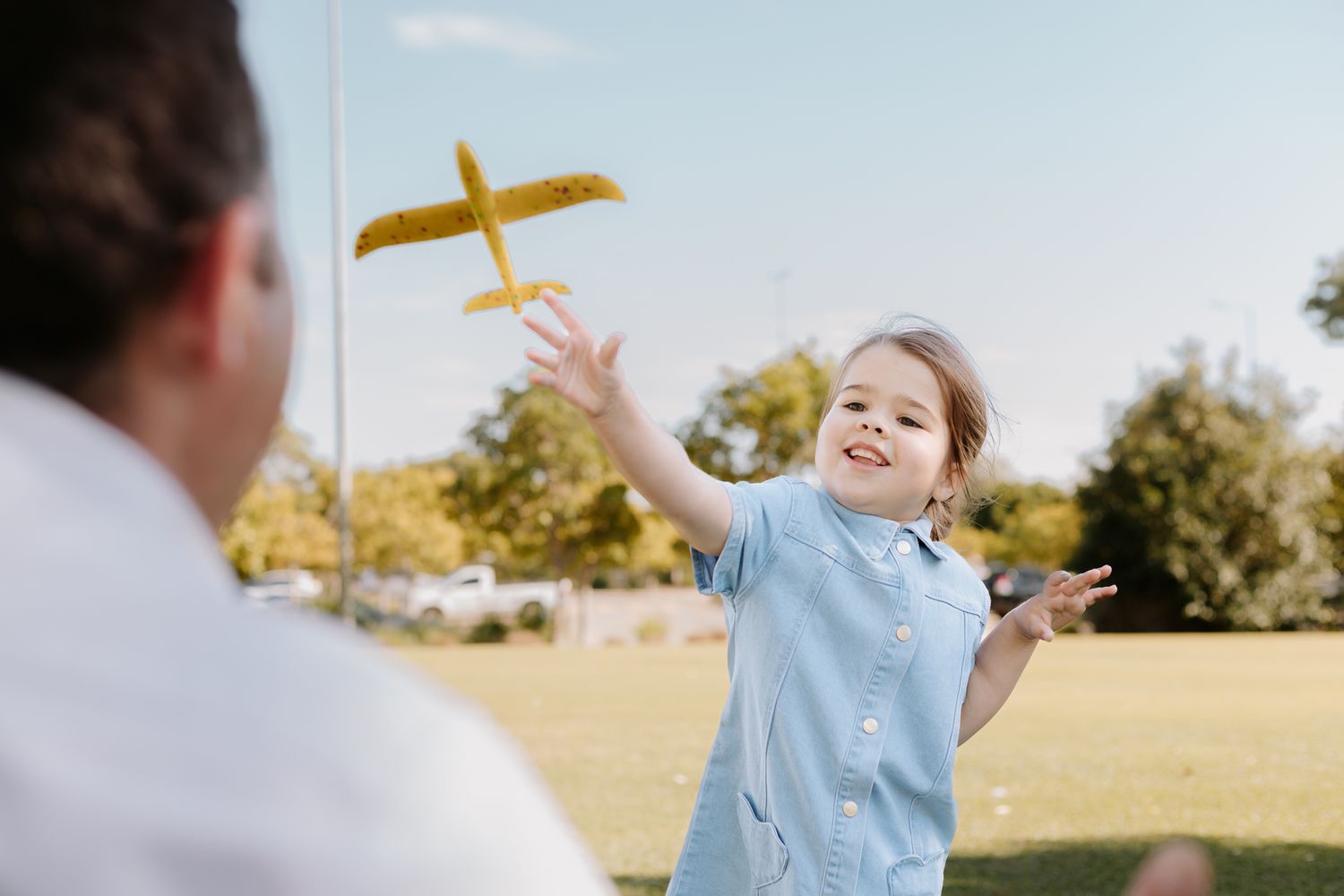 A happy child tosses a yellow toy airplane in a sunny park while playing with an adult outdoors.
