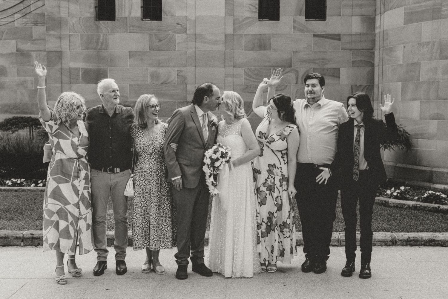 Group photo of wedding party lined up against stone building wall in formal attire.