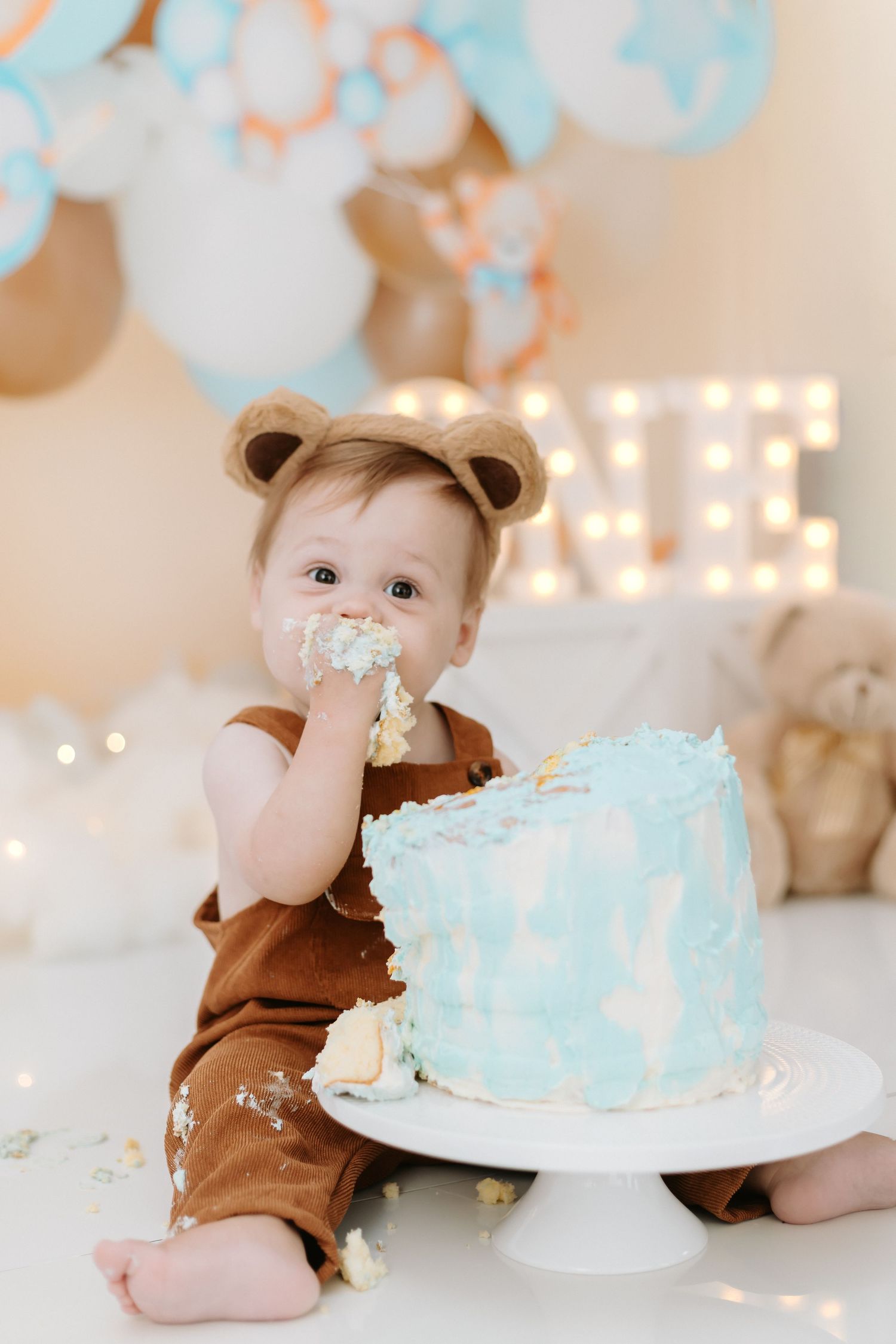 Baby in brown bear costume having first birthday cake smash photoshoot with blue cake and twinkle lights.