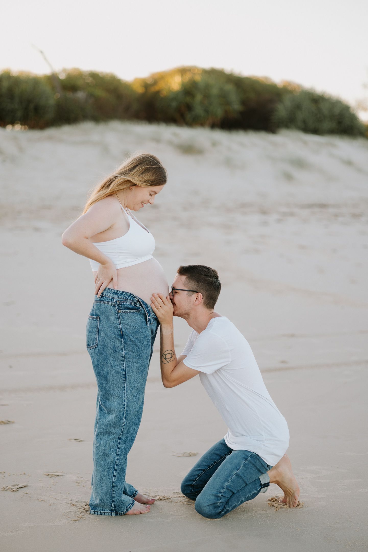 A couple shares an intimate moment on a sandy beach at sunset with soft natural lighting.