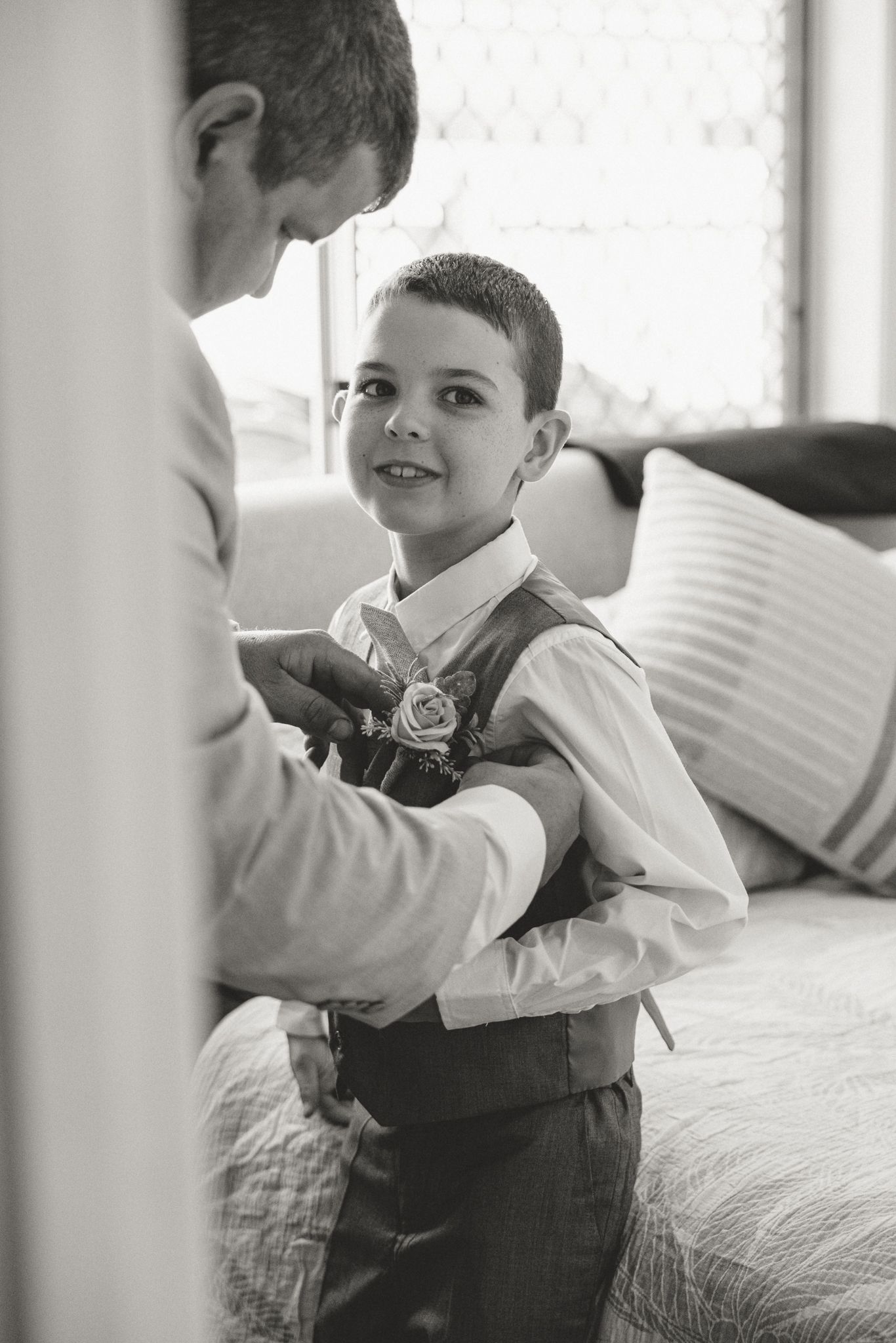 Black and white photo of a man pinning a boutonniere on a smiling young boy in a vest at a wedding.