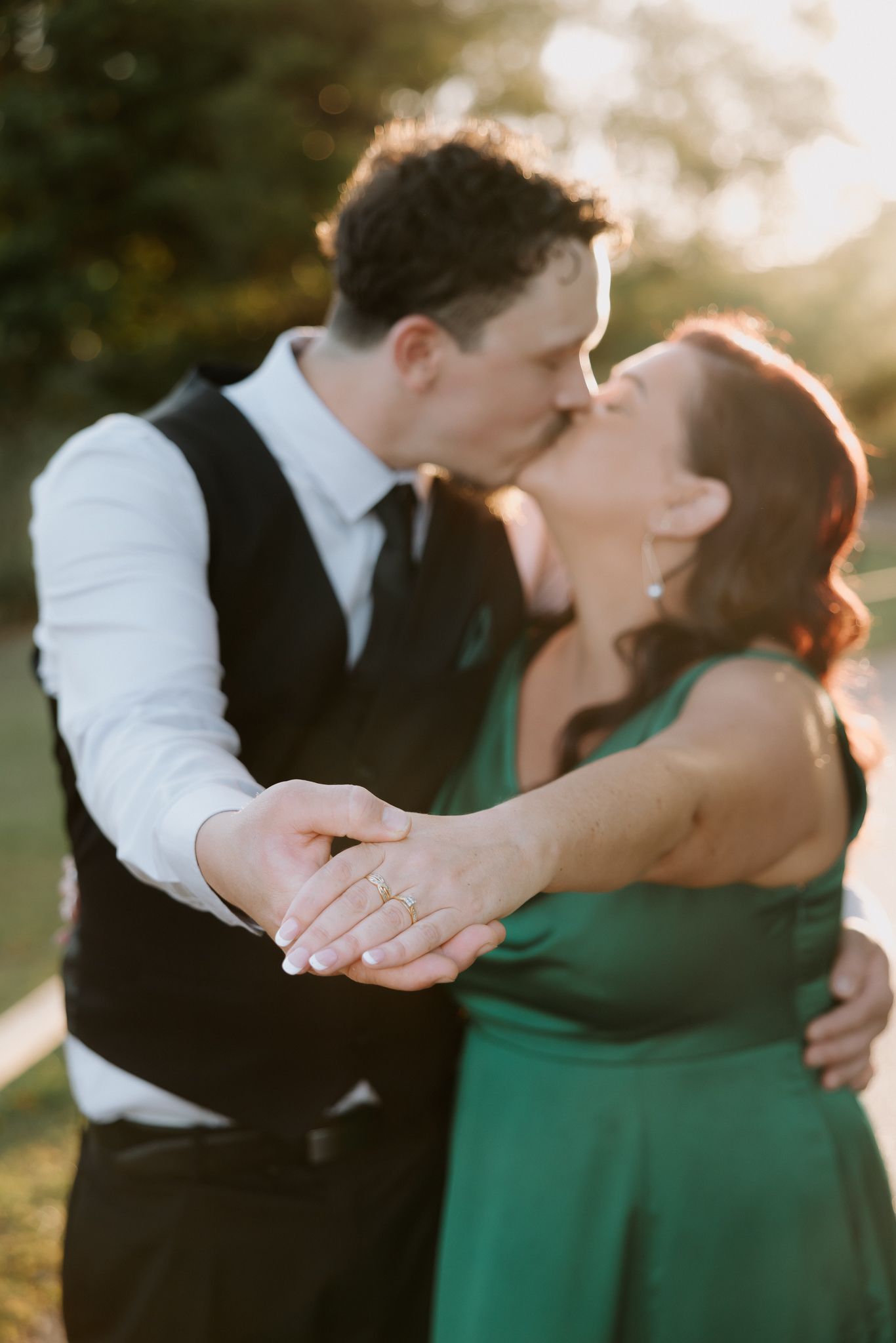 Couple sharing a romantic kiss at golden hour, woman in green dress showing engagement ring while embraced outdoors.