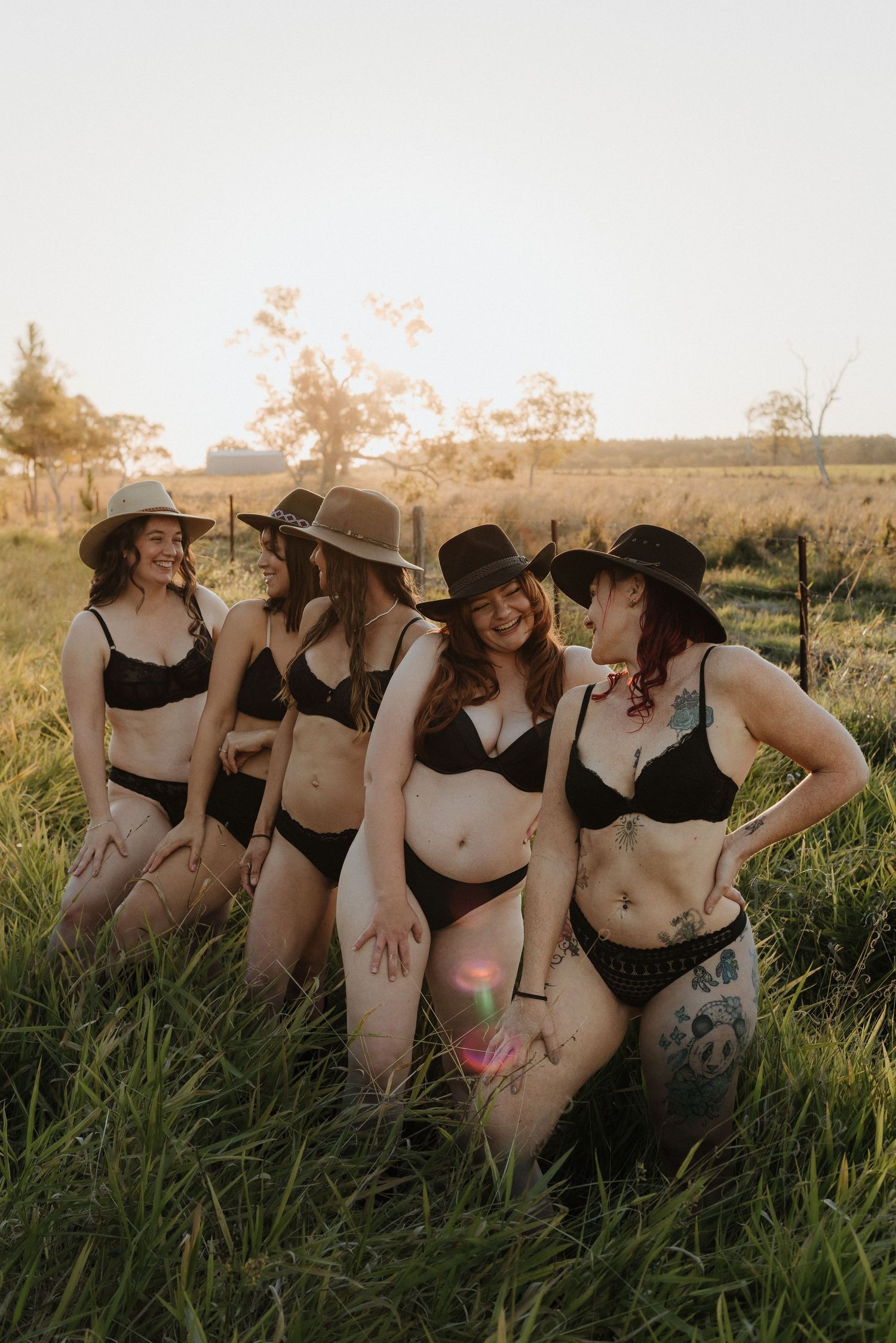 Four women in matching black lingerie and wide-brim hats pose together in a sunlit green field at golden hour.
