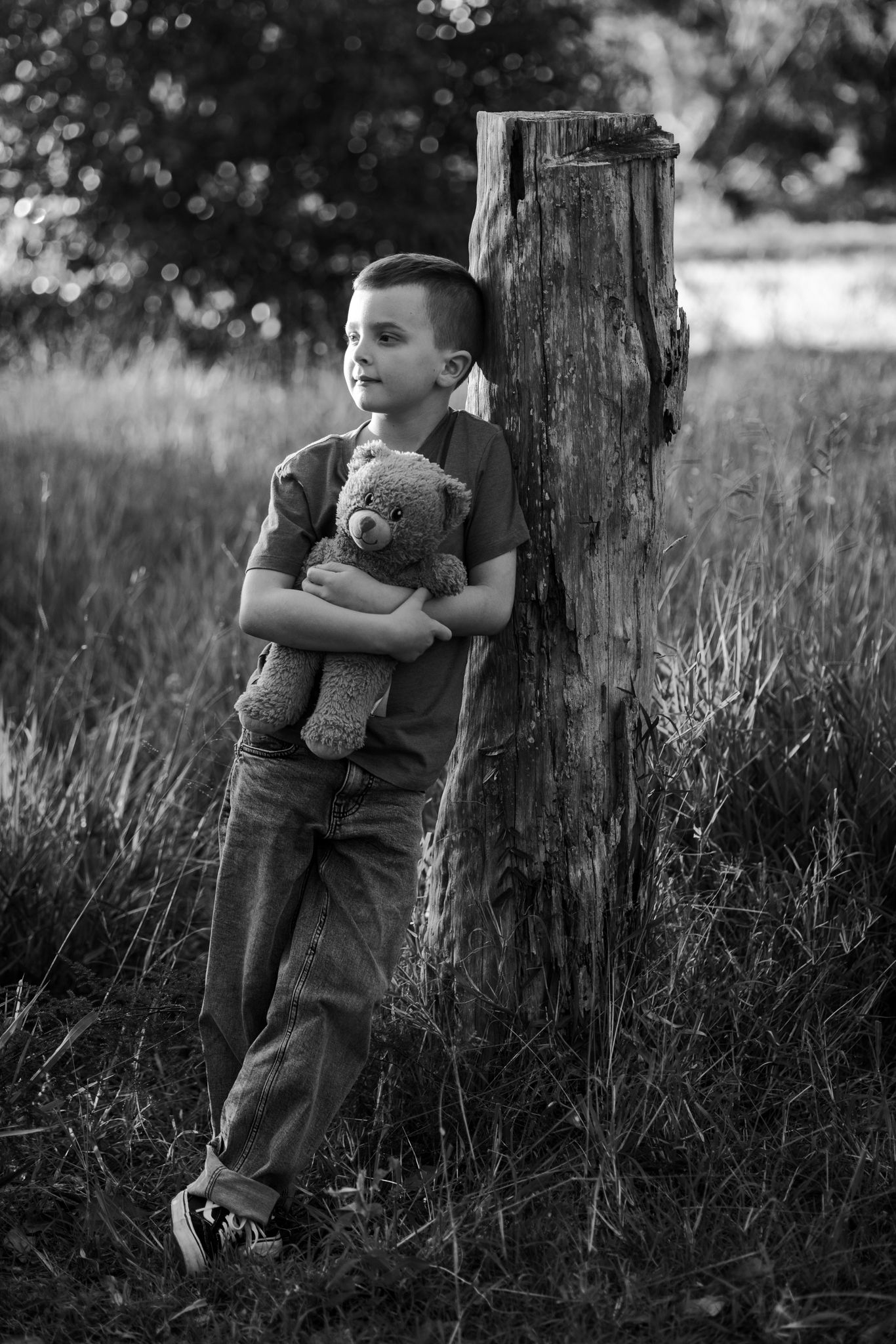 Black and white photo of a young boy leaning against a wooden post outdoors, hugging a teddy bear.