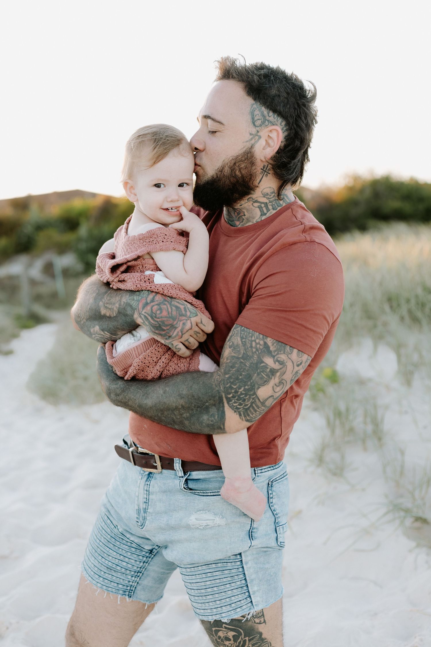 A tattooed person in a coral shirt and denim shorts holds a baby on a sandy beach at sunset.