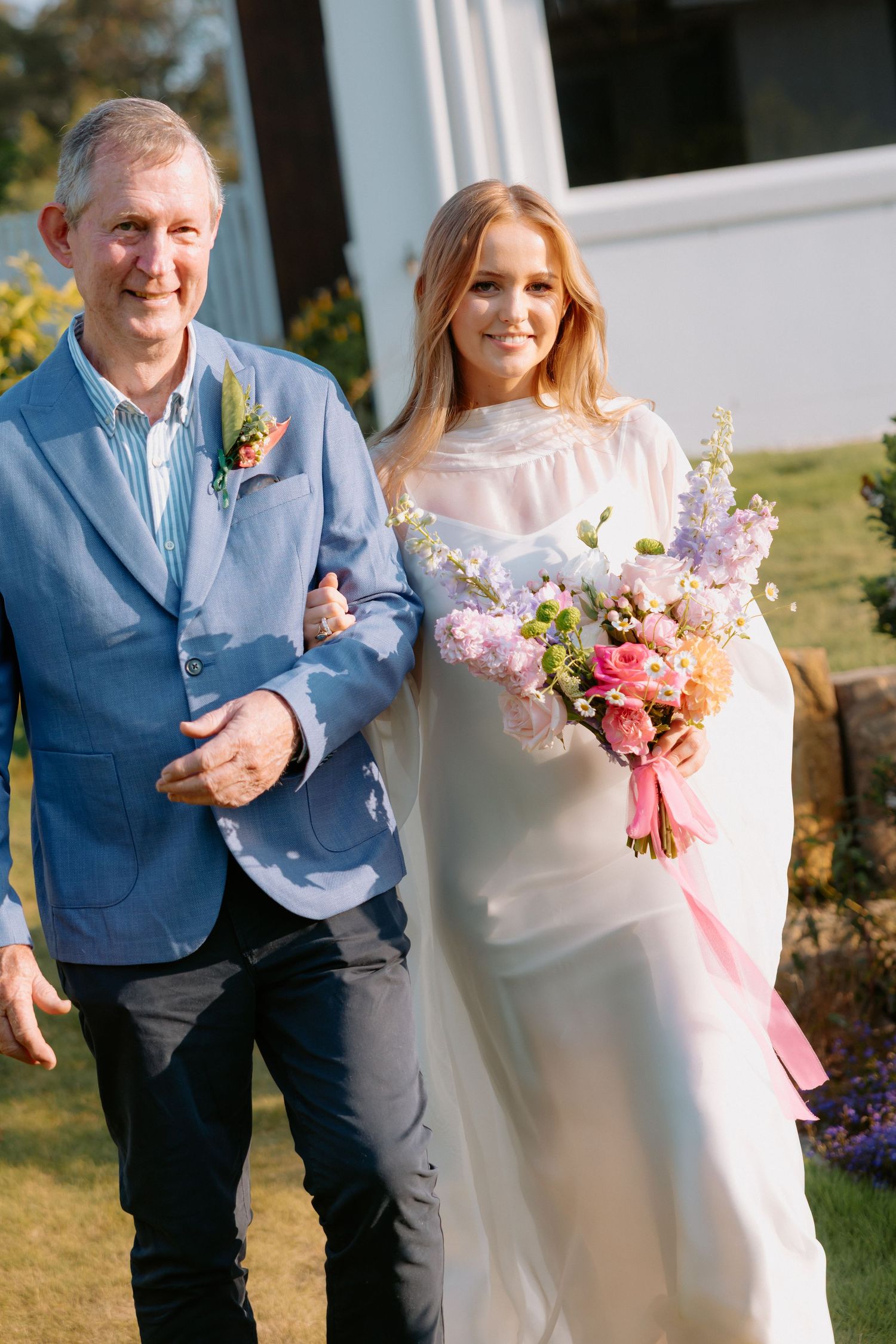 Two people in formal attire share a joyful moment outdoors with floral bouquets.