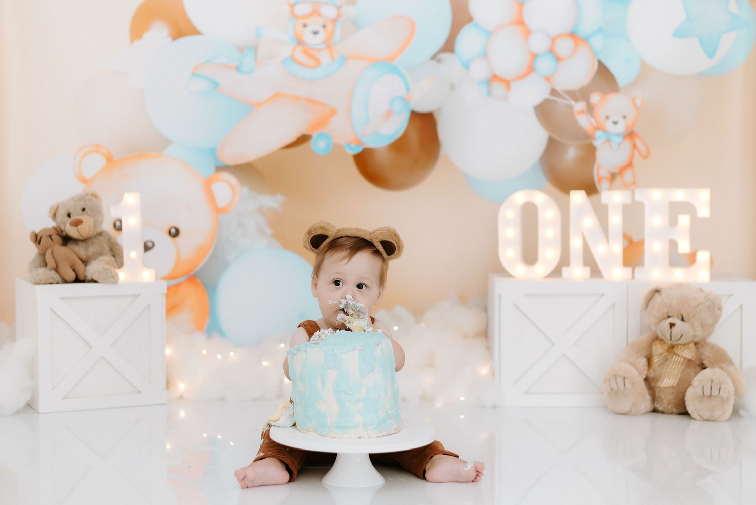 First birthday celebration setup with pastel blue and peach balloons, white boxes, teddy bears, and a small cake.