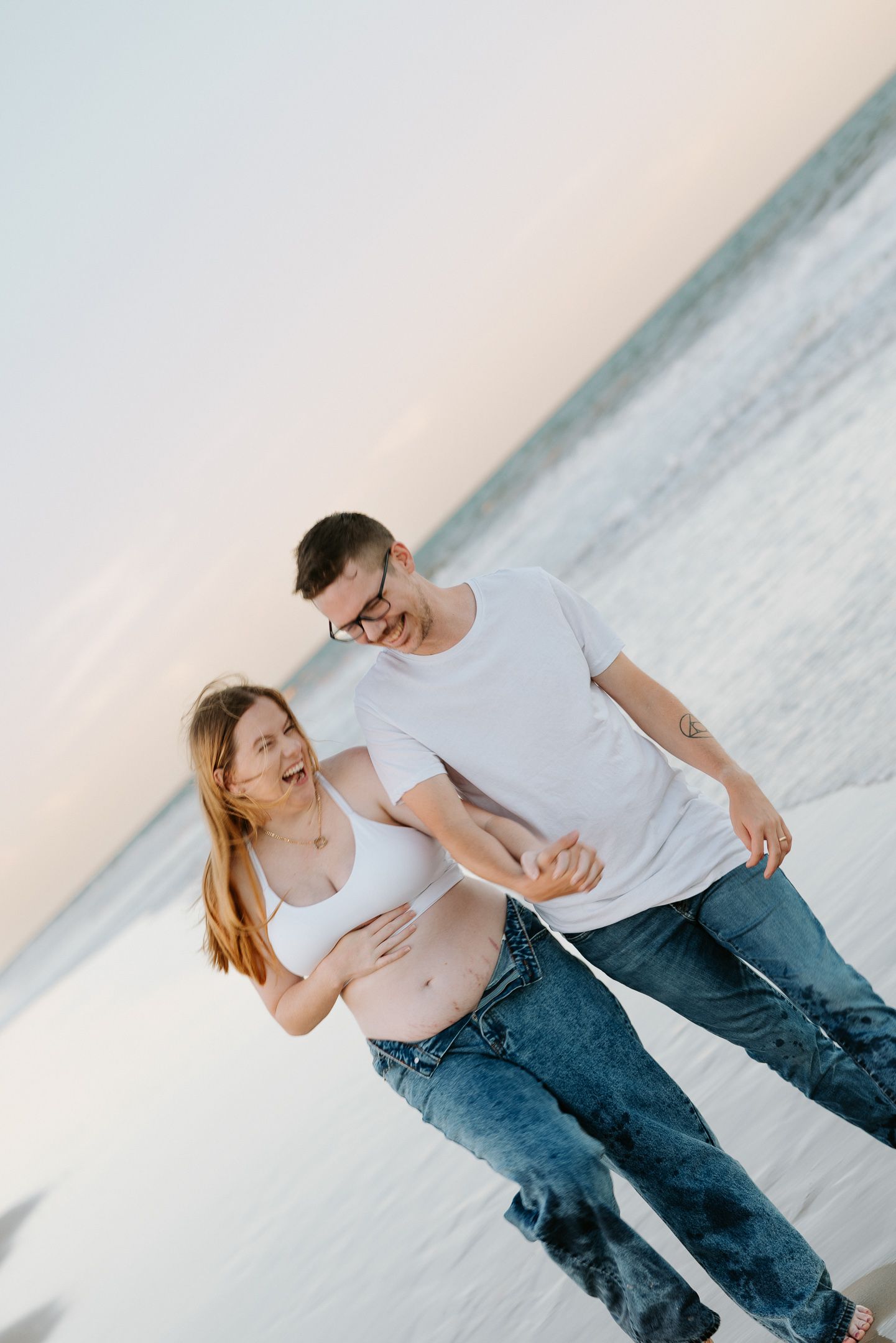 A couple in white shirts and jeans shares a playful moment together on a cloudy beach.