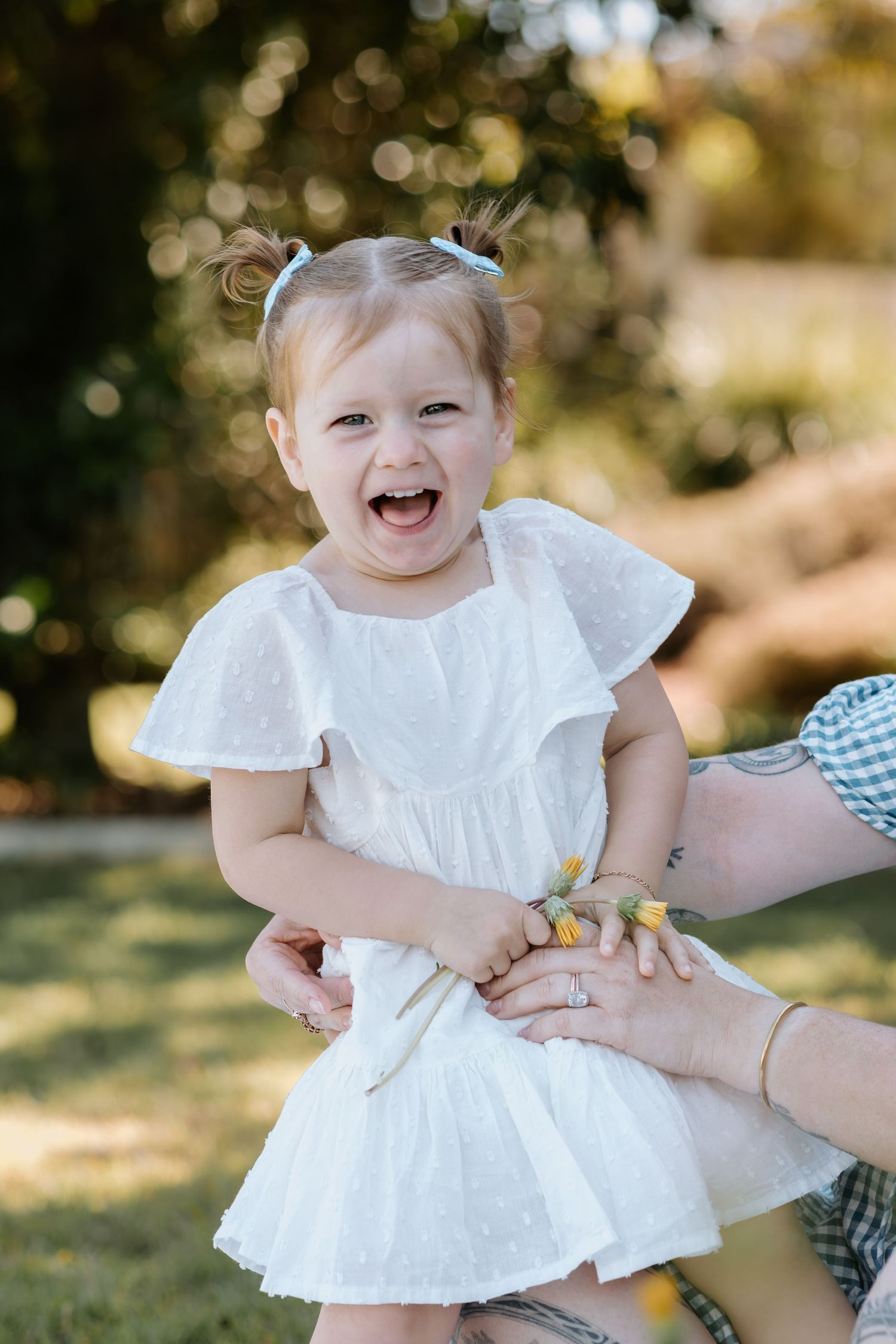 A joyful toddler girl in a white dress laughs outdoors while being held by an adult in a sunny garden setting.