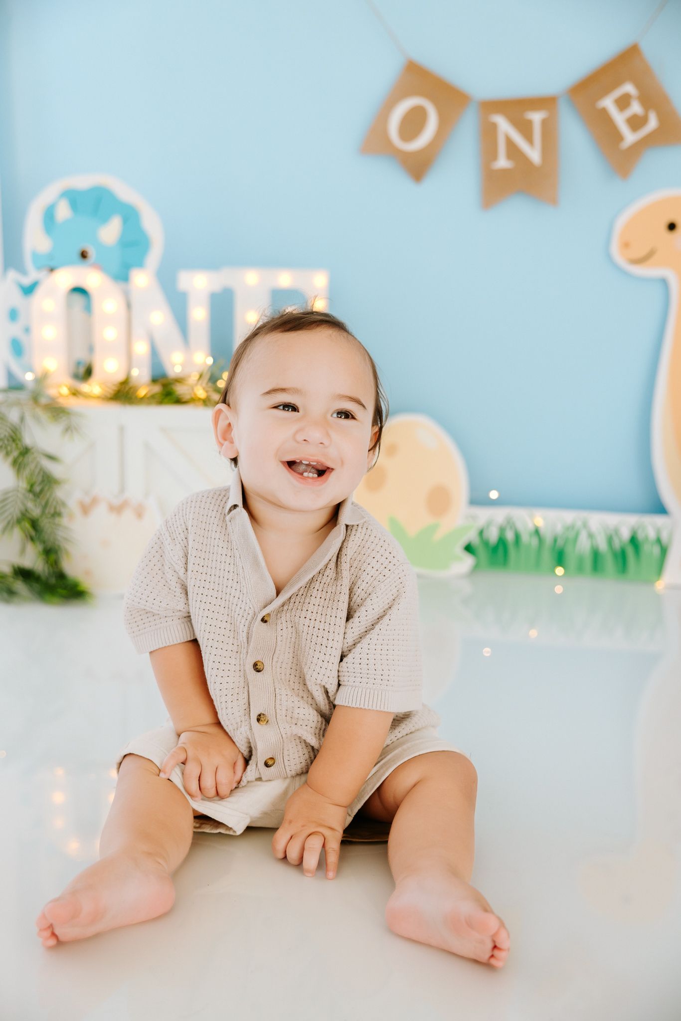 Smiling baby boy sitting on floor at first birthday party with ONE banner and dinosaur decorations.