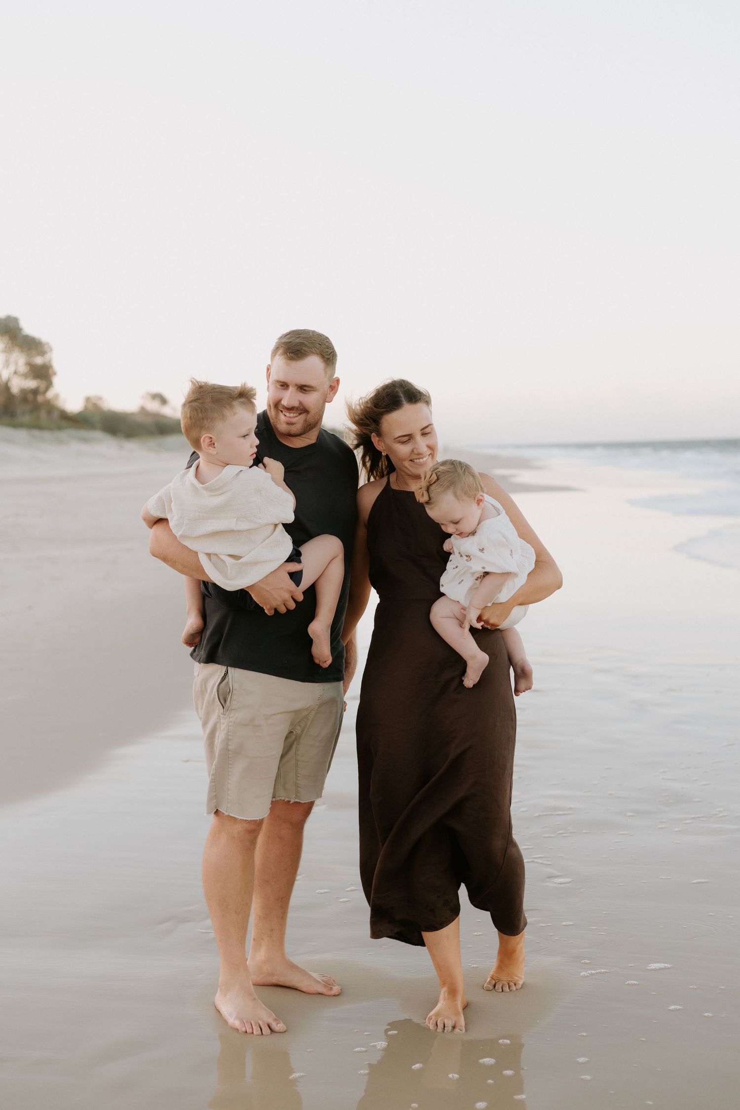 Family of four smiling on a sandy beach at sunset, parents holding two young children, barefoot in shallow water.
