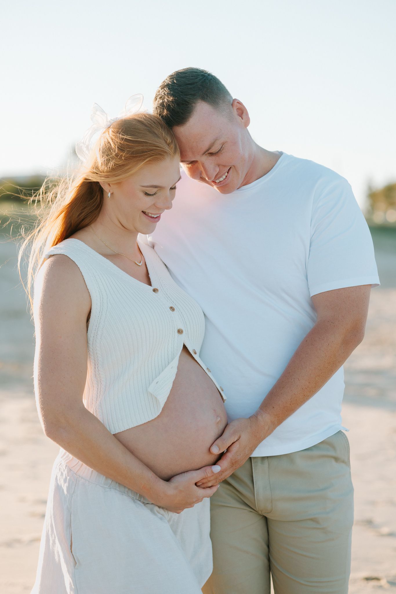 A happy couple cradles a pregnant belly on a sunny beach, the woman in white dress with red hair, man in white tee.