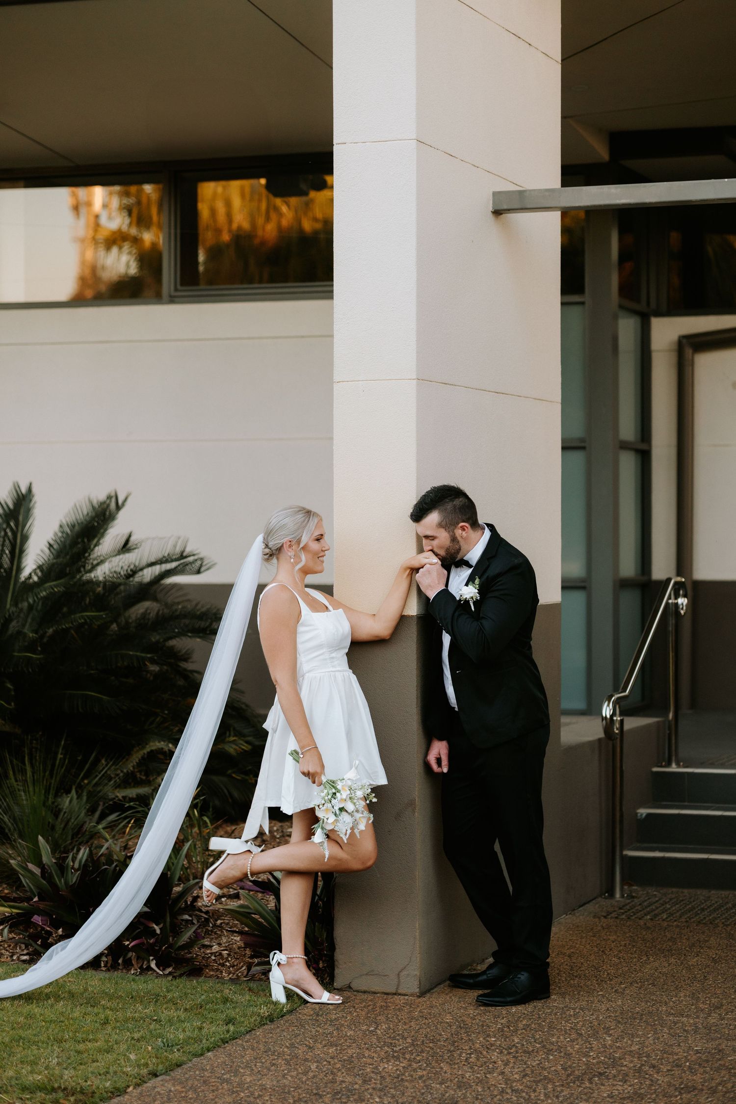 Groom in black tuxedo kisses bride's hand as she leans against a pillar in a short white dress with long veil.