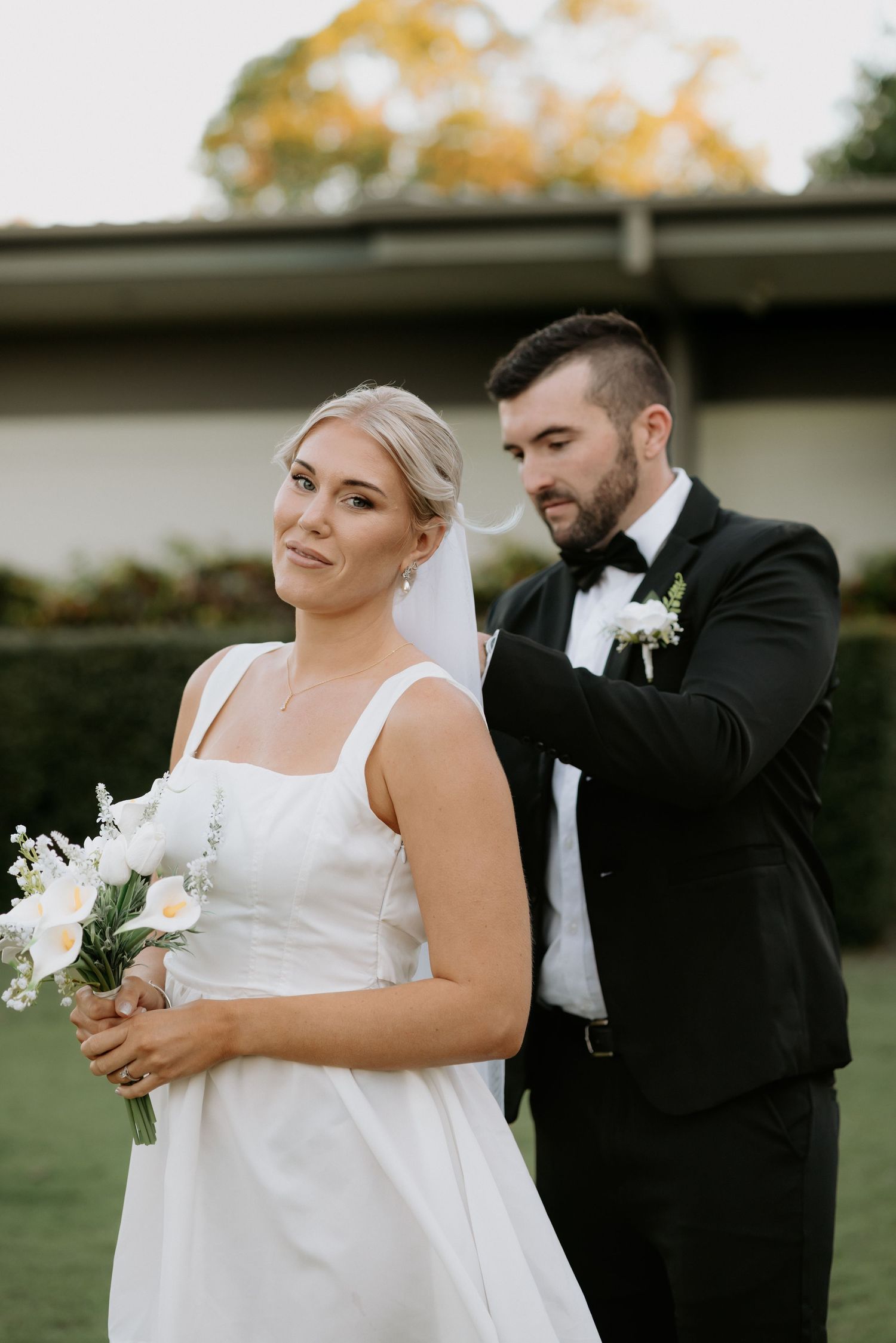Groom in black tuxedo adjusting bride's veil outdoors, bride holding white floral bouquet in elegant wedding dress.