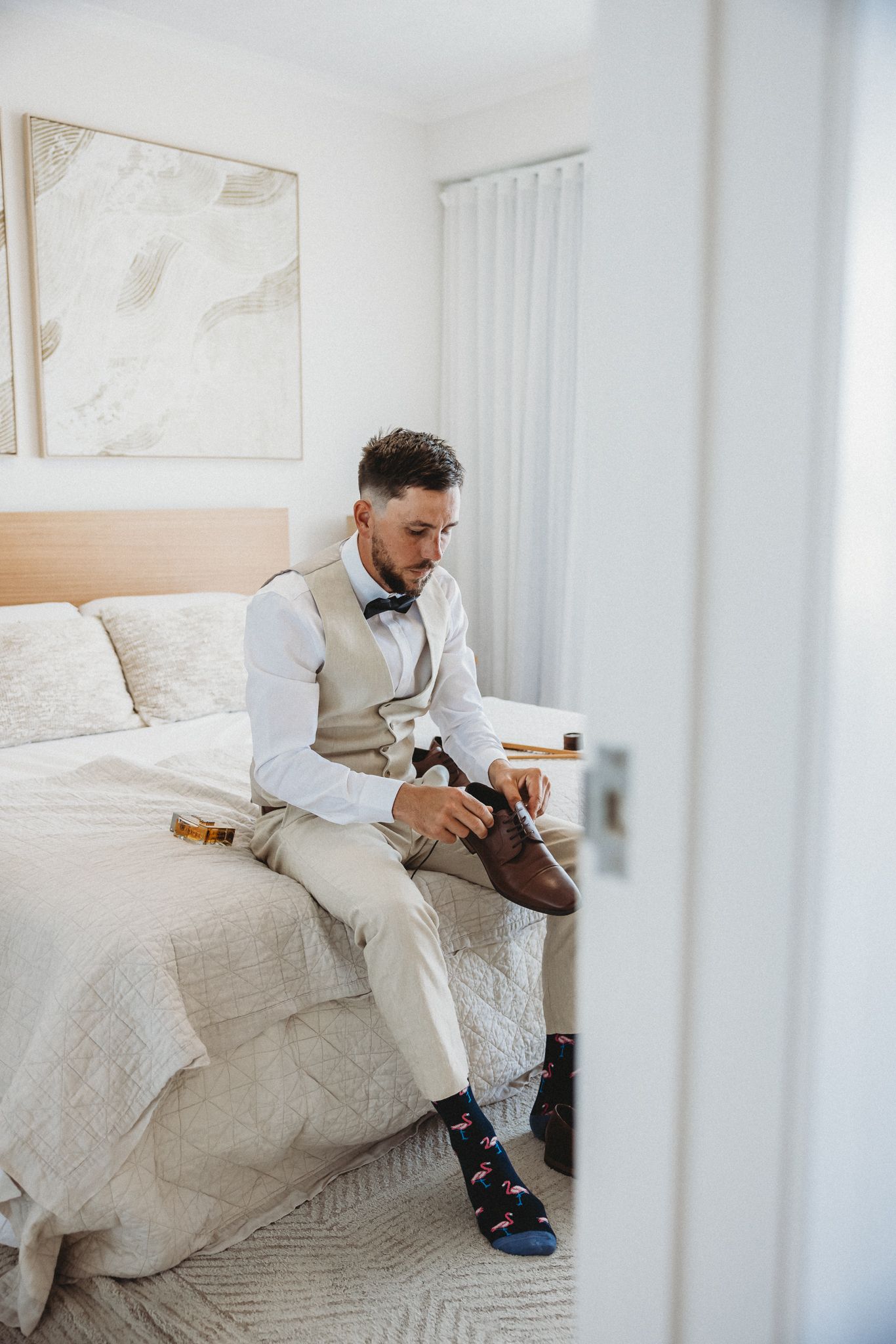 Groom in beige vest and bow tie sitting on bed putting on brown dress shoes on wedding day.