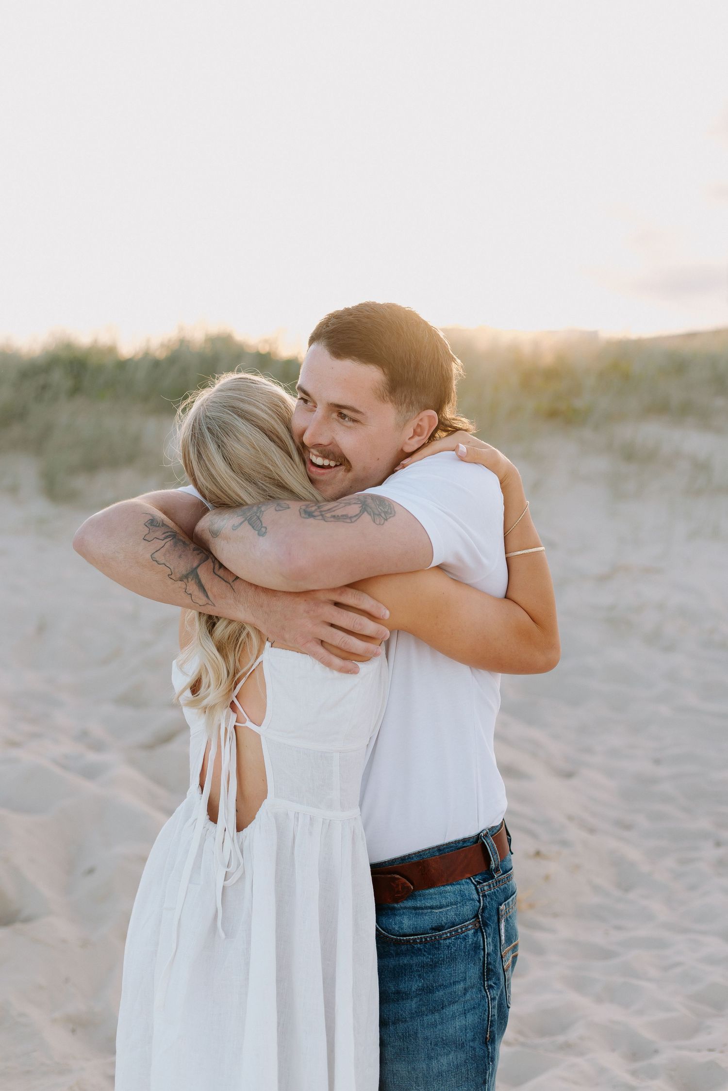 A smiling couple embraces on a sandy beach at sunset, man with tattoos in jeans and white tee hugging woman in white dress.